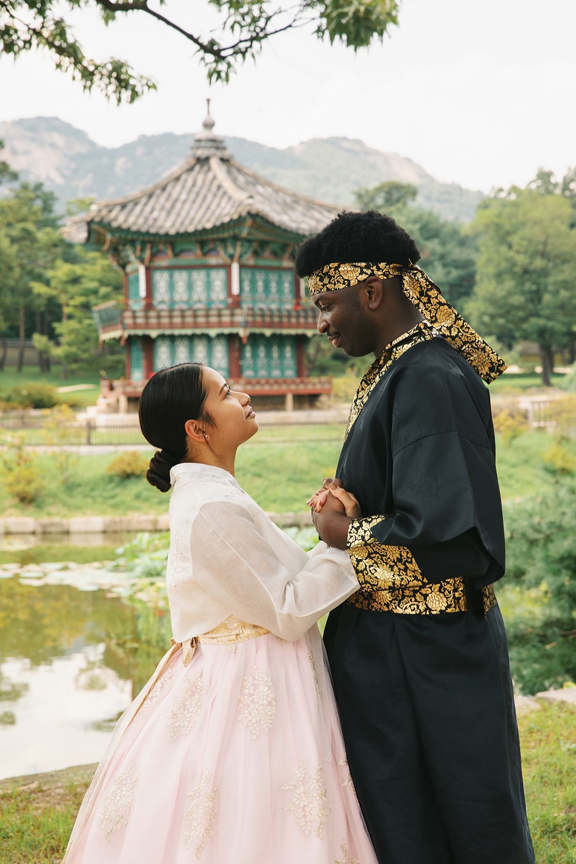 A couple wearing traditional Korean hanbok attire shares a playful moment near a traditional pavilion by a lake.