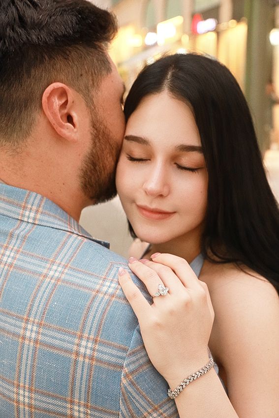 A romantic couple embraces while showing off an engagement ring in a soft glowing light.