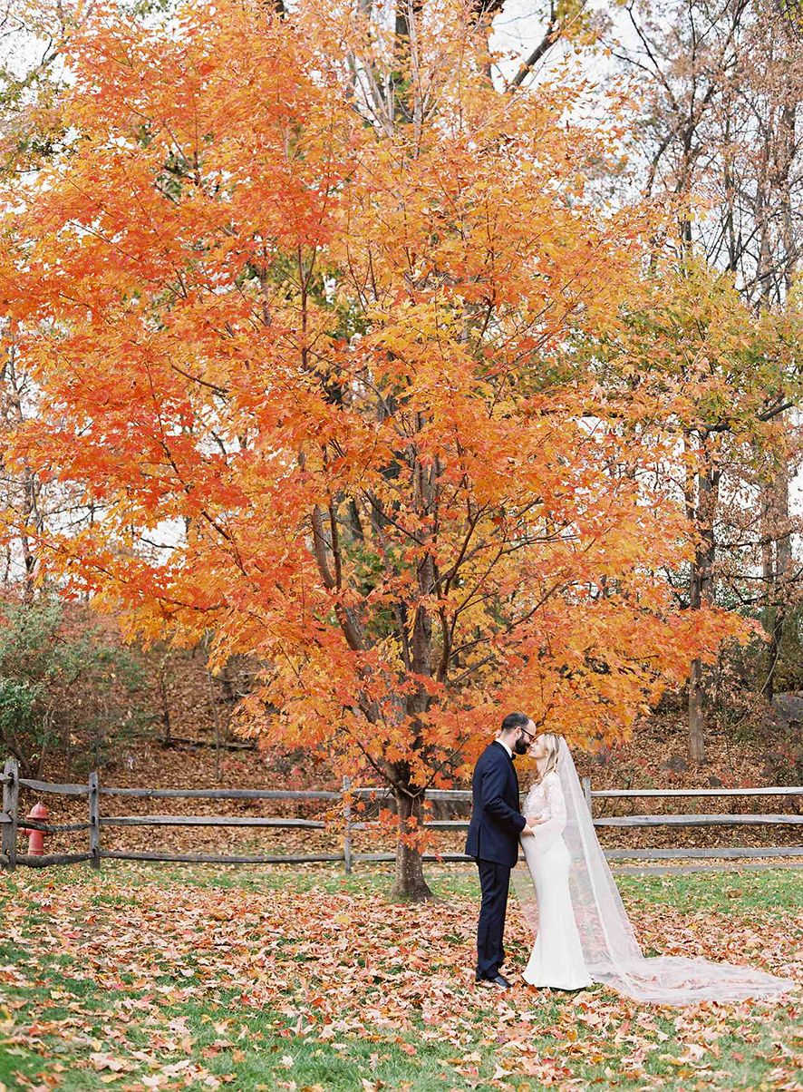 A couple stands beneath a vibrant orange maple tree during their autumn wedding with fallen leaves scattered on the ground.