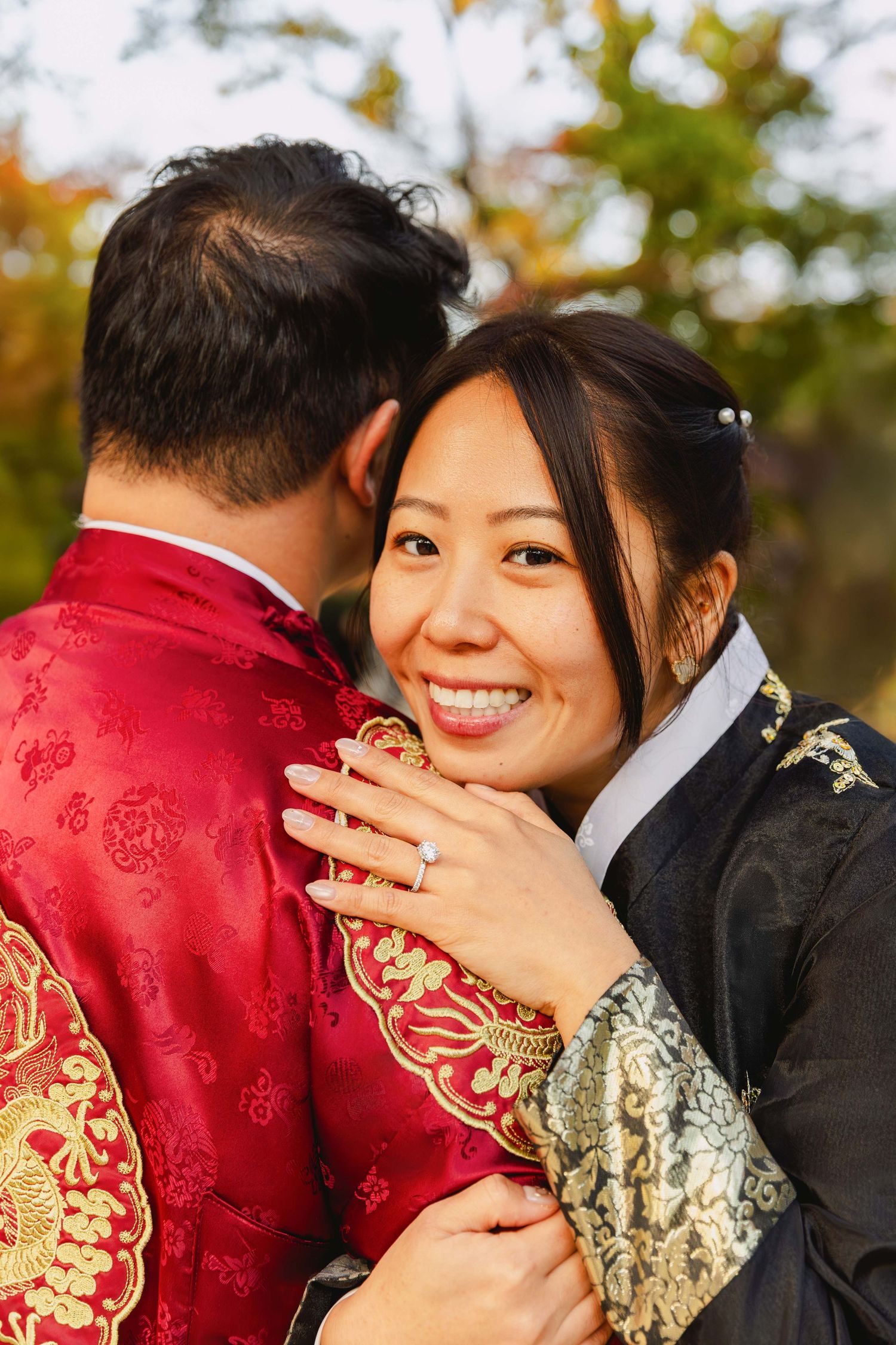 Smiling Asian bride in traditional attire embraces groom in red Chinese wedding outfit outdoors.