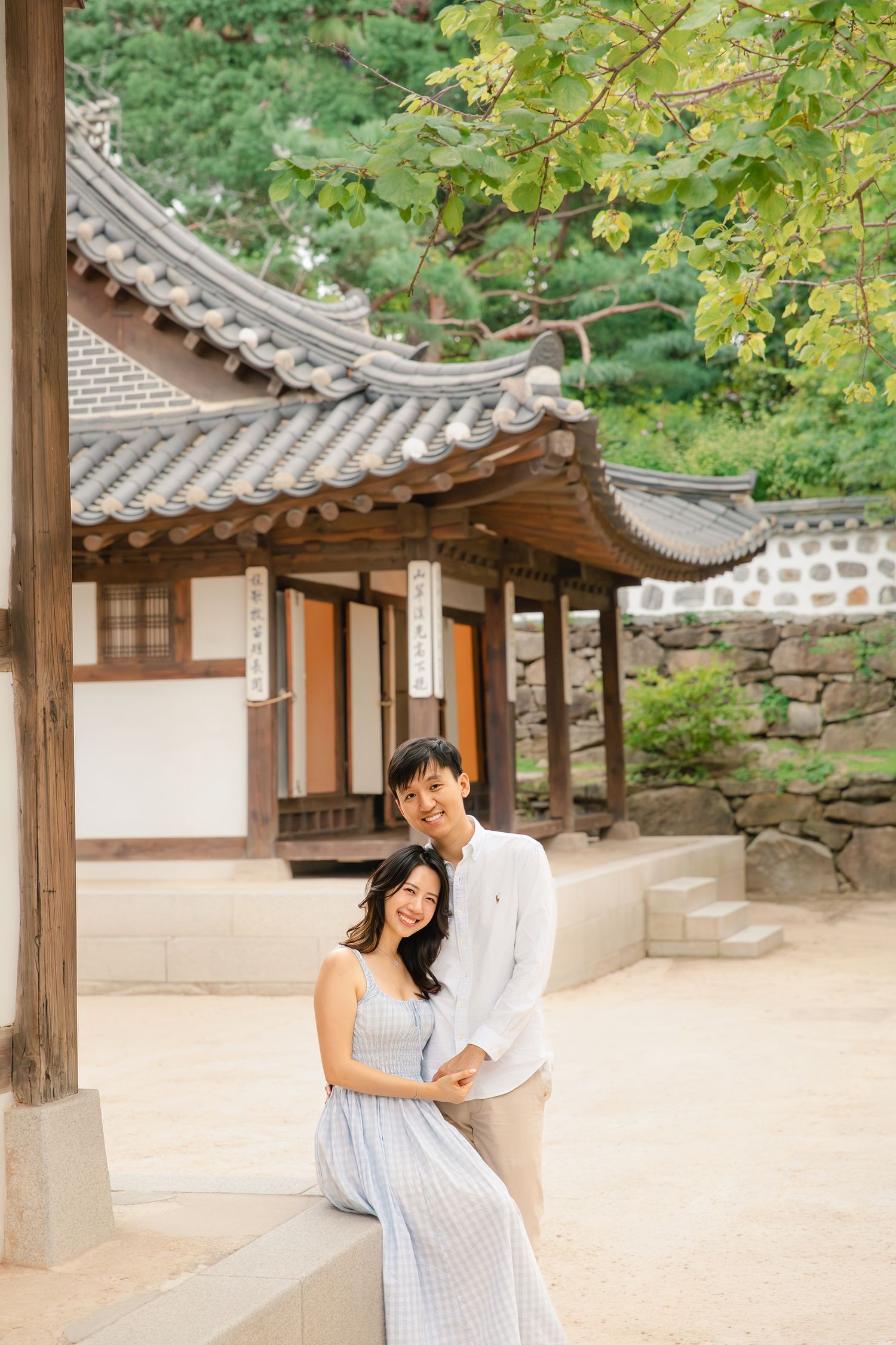 Traditional Korean hanok building with curved tile roof and wooden walkway surrounded by lush green foliage.