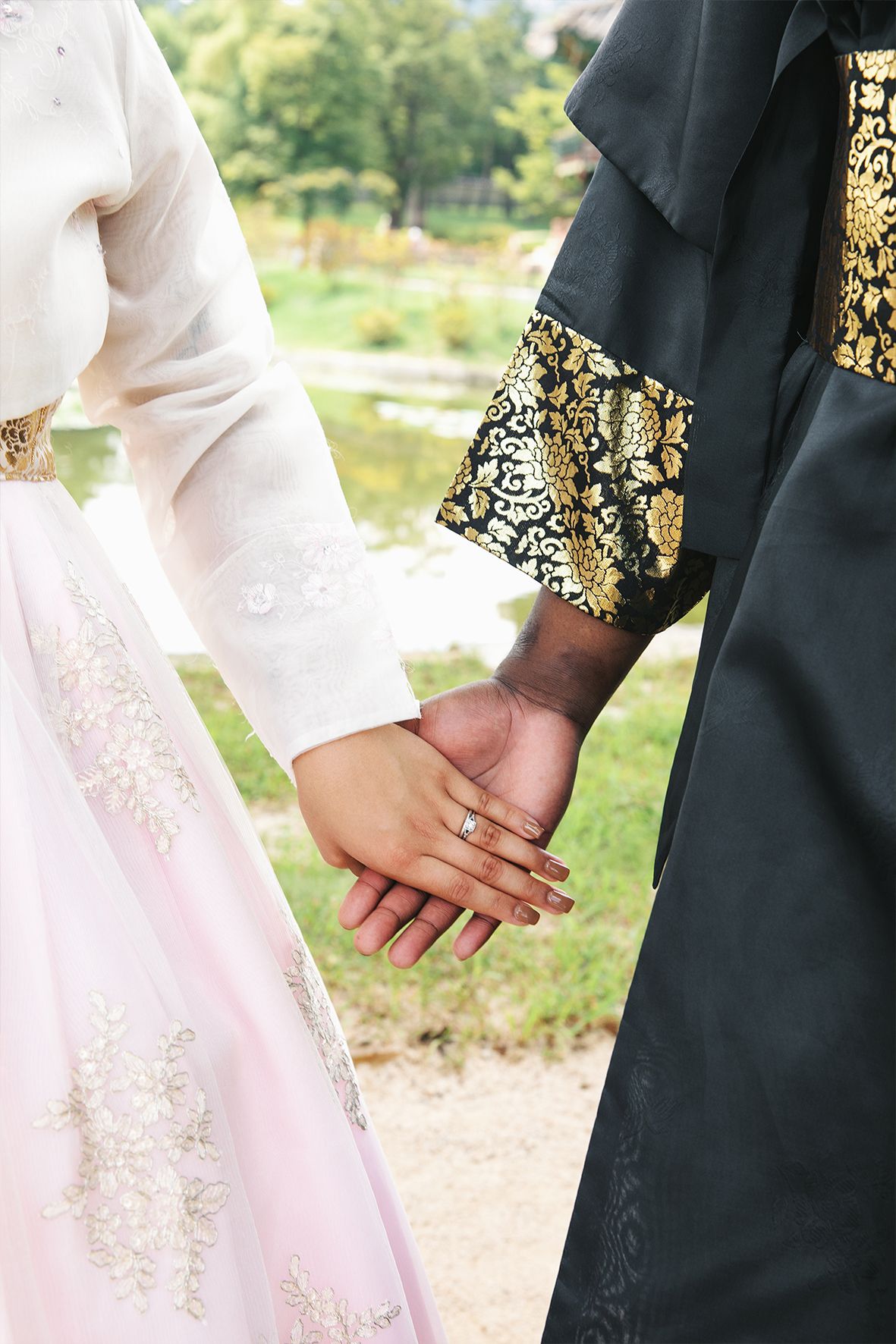 Couple in traditional Korean hanbok holding hands, with white and black silk robes adorned in gold embroidery details.