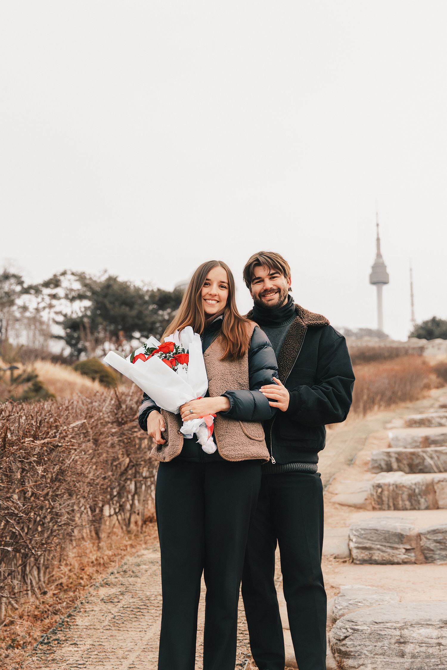 A couple in matching black outfits stands together on a winding path during autumn with a tower in the distance.