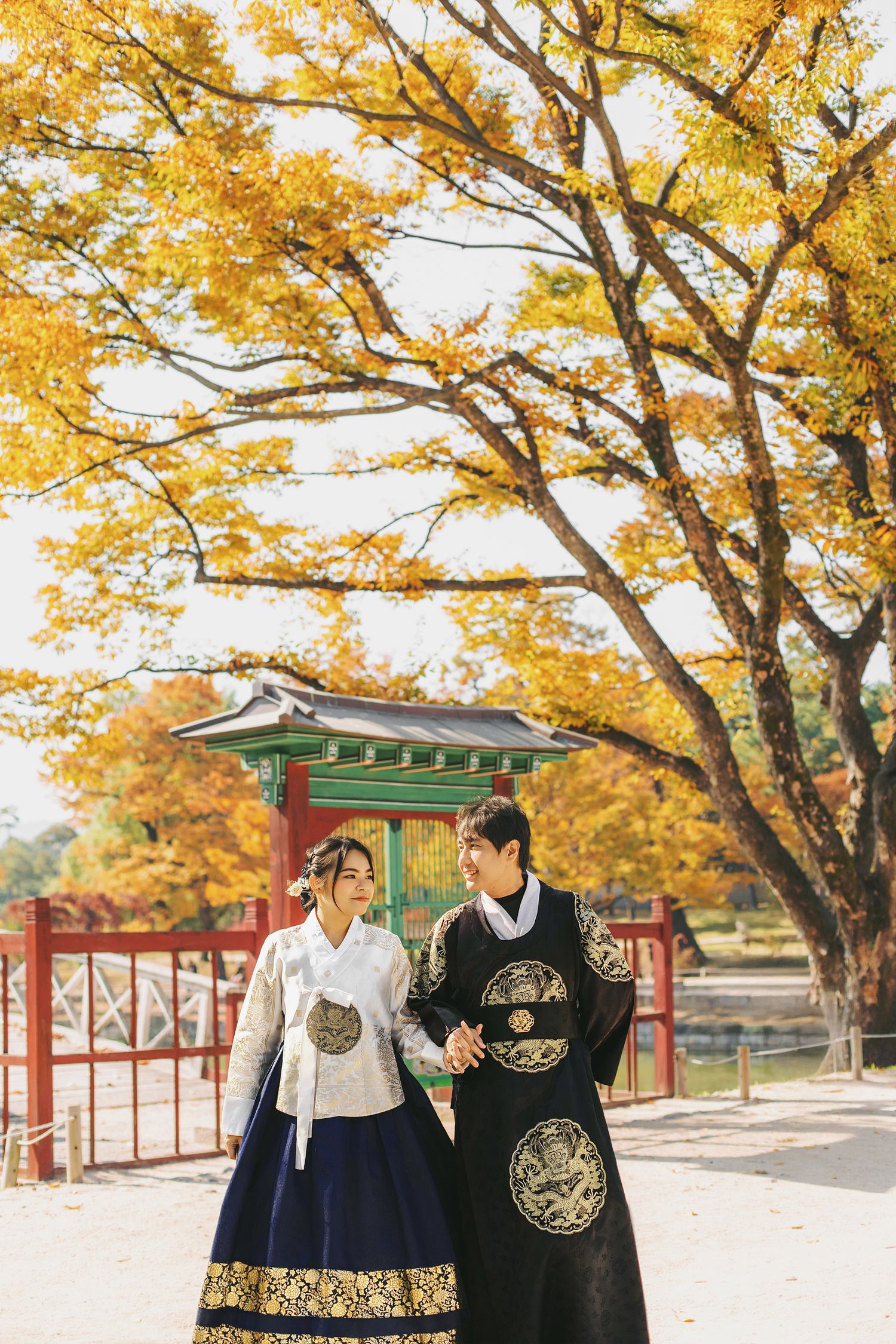 Couple wearing traditional Korean hanbok outfits walking beneath golden autumn trees in a historic palace courtyard.