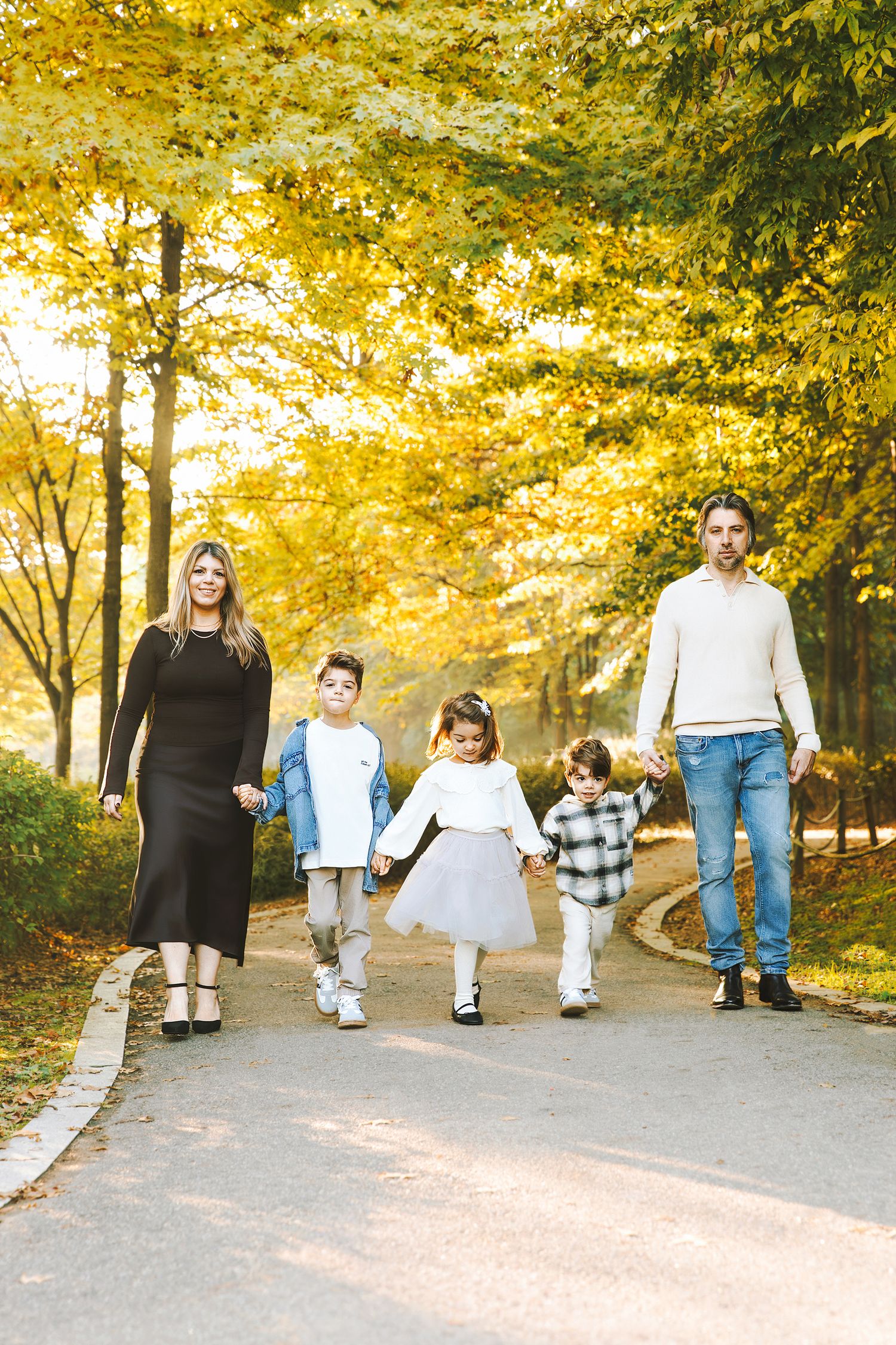 Family of five walking hand-in-hand on a tree-lined path surrounded by golden autumn foliage.