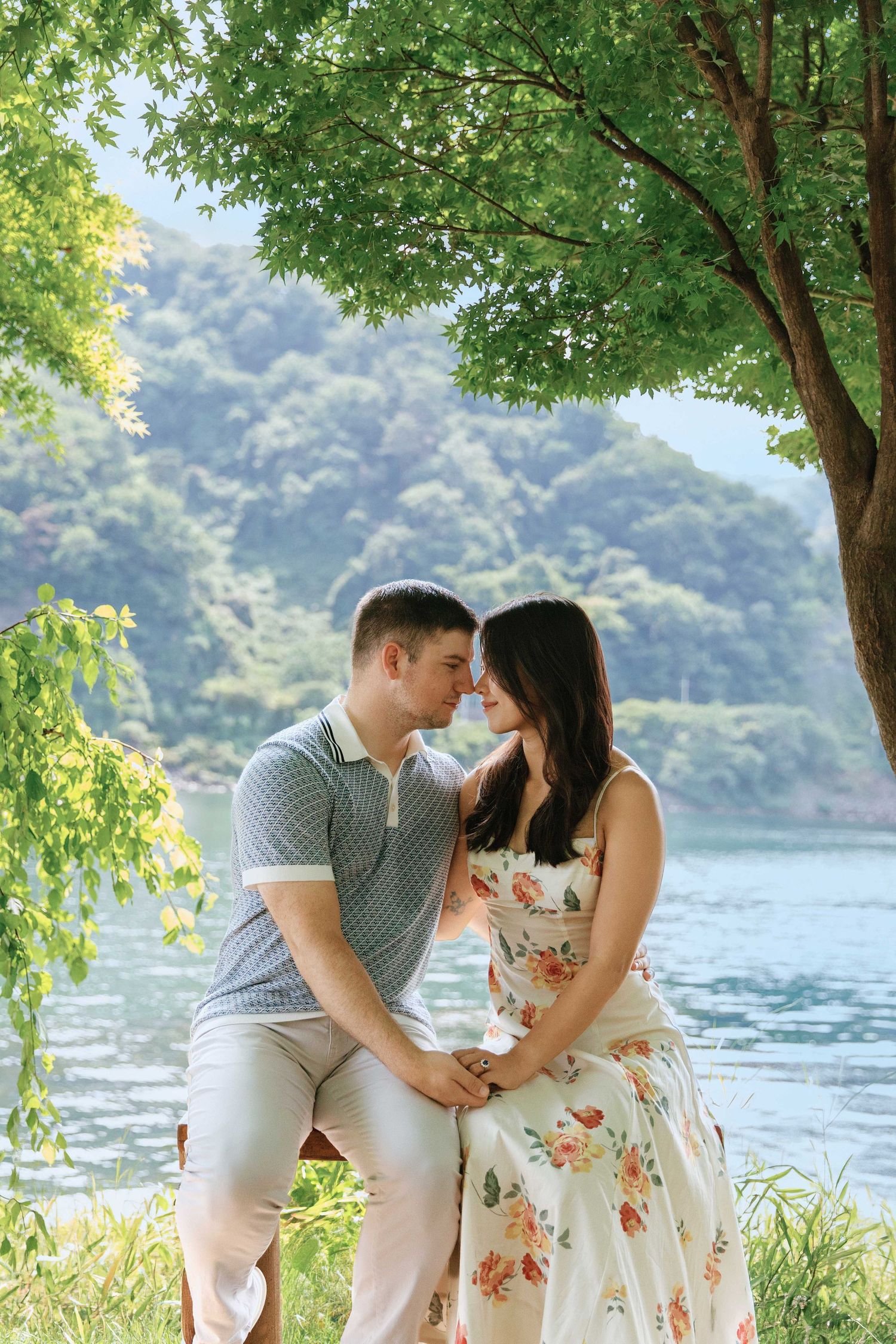 Romantic couple sitting close together under a lush green tree by a serene lake, surrounded by misty mountains.