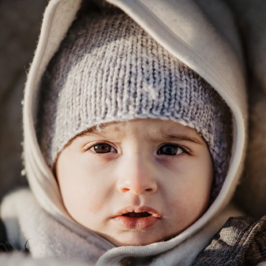 A child wearing a knit winter hat and cream colored hood looks at the camera with rosy cheeks.