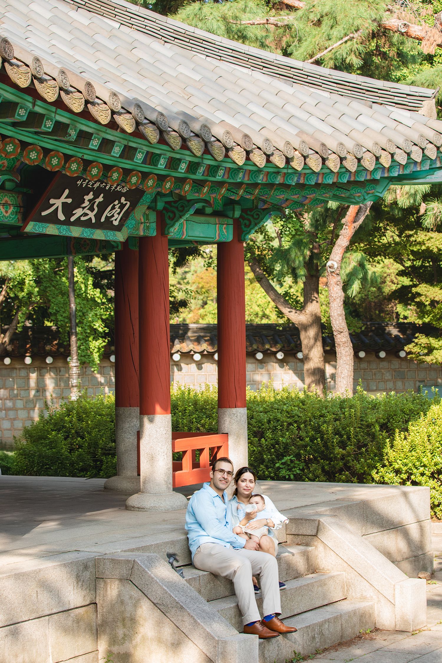 Traditional Korean covered walkway with red pillars and ornate tiled roof in a peaceful garden setting.