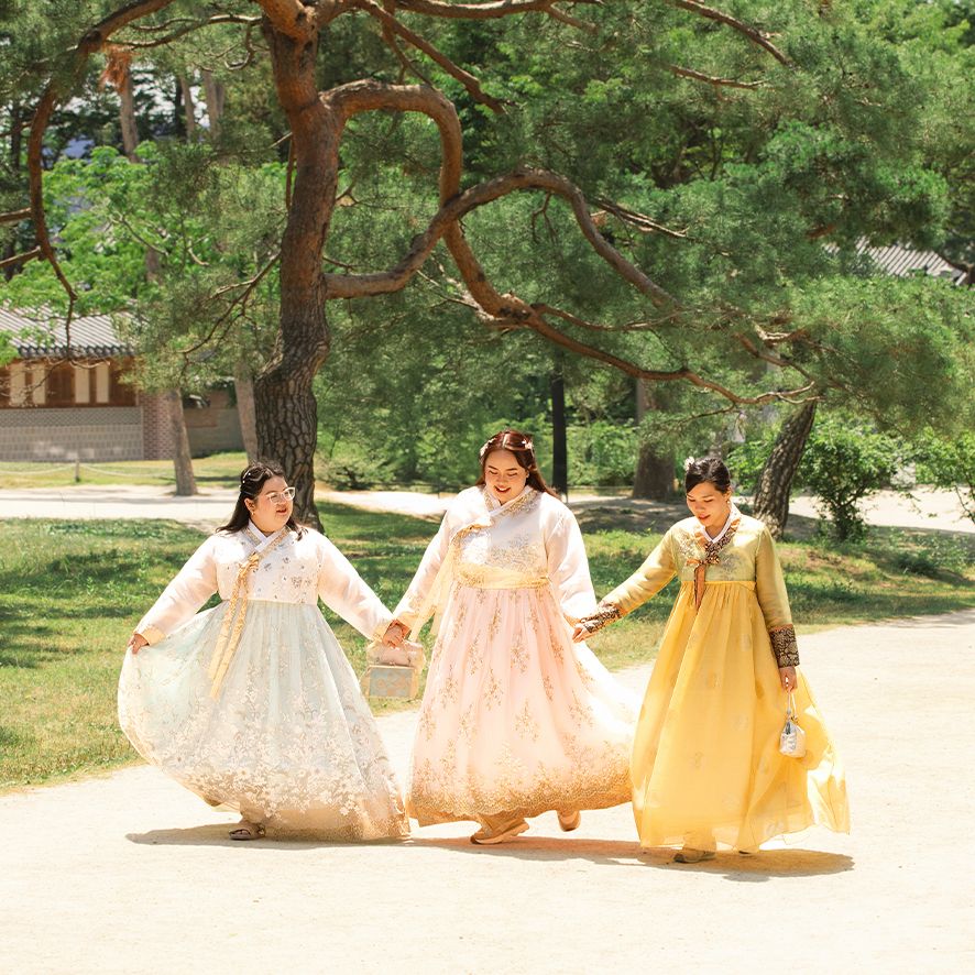 Three friends in traditional Korean hanbok dresses hold hands while walking together in a sunny park with pine trees.
