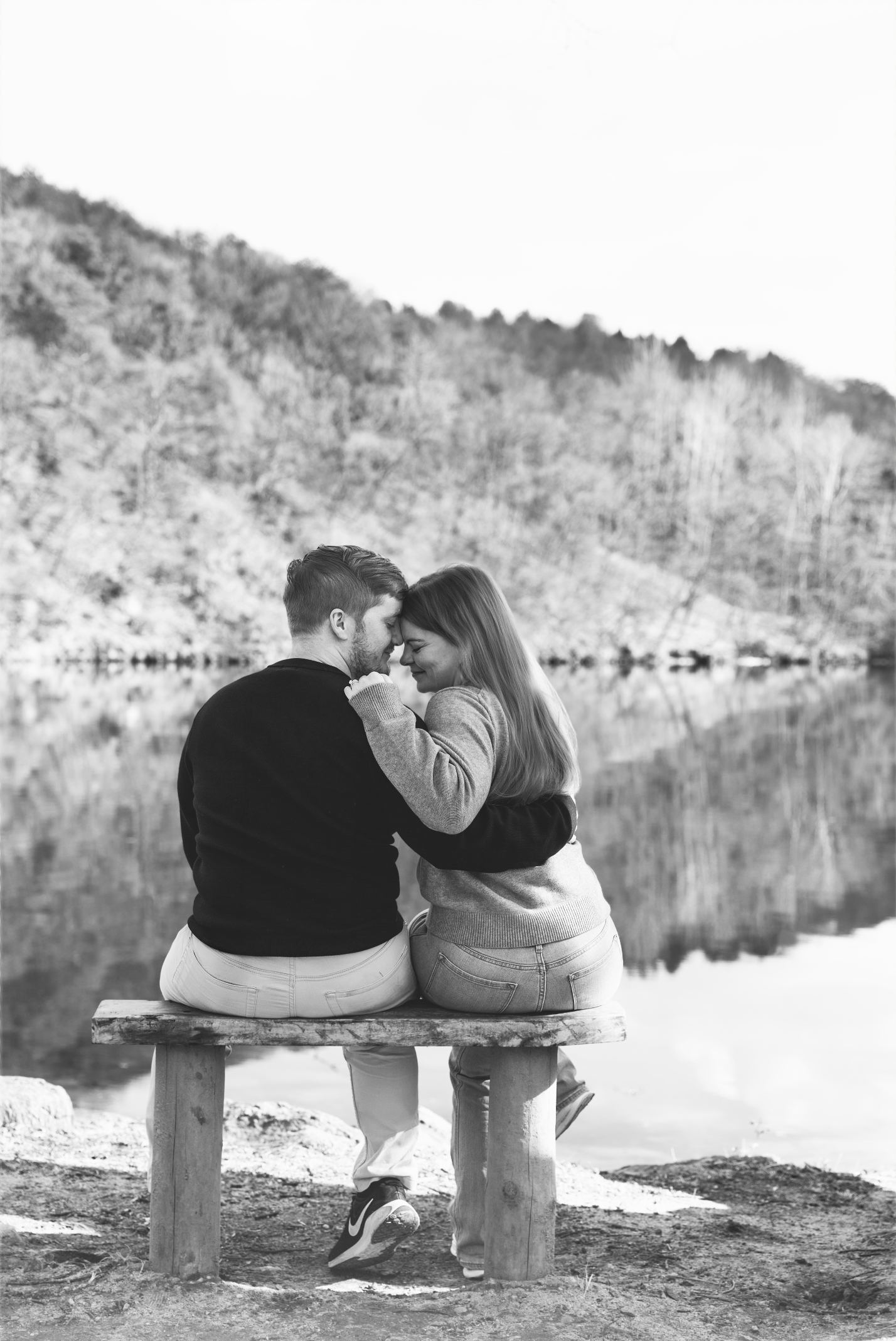 A romantic couple sits together on a wooden bench by a serene lake with mountains reflecting in the still water.