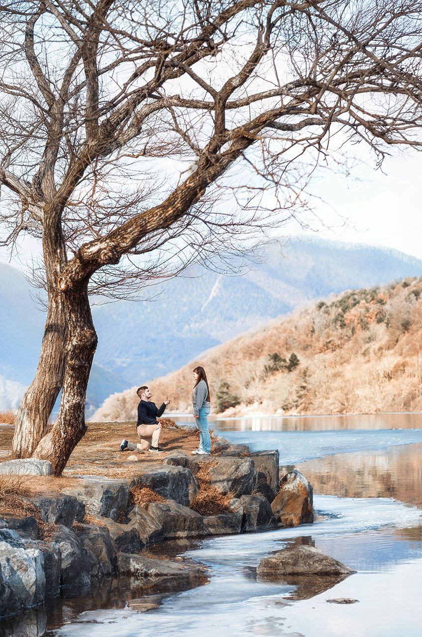 People sitting by a serene lake surrounded by mountains and bare winter trees in a peaceful outdoor setting.