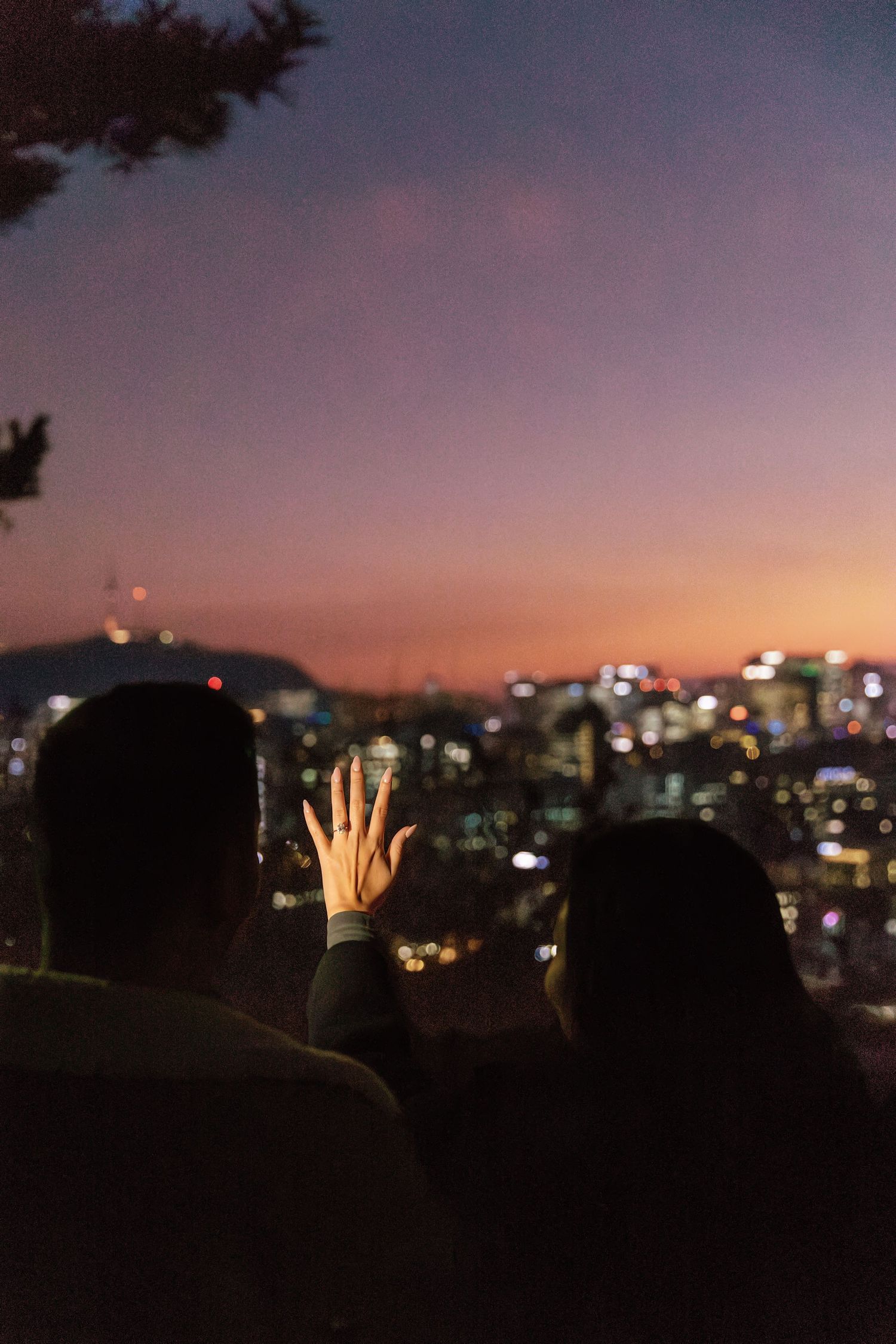 Couple enjoying a romantic city skyline view at dusk, with bokeh city lights and a colorful purple-pink sunset sky.