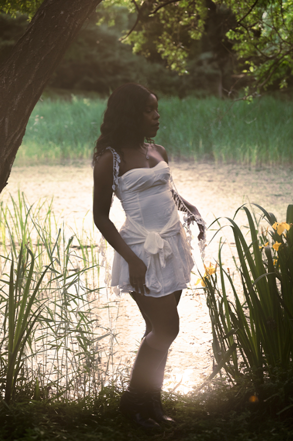 A silhouetted figure in a white summer dress poses against backlit foliage in a dreamy outdoor setting.