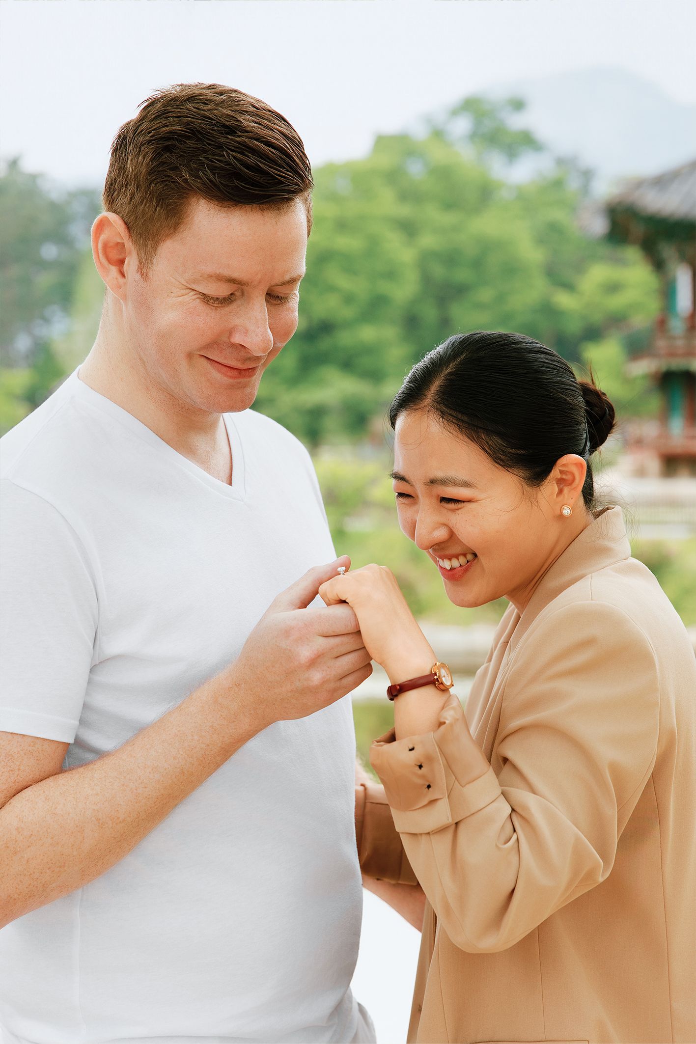 Smiling interracial couple sharing a tender moment outdoors near a Korean pavilion surrounded by lush greenery.