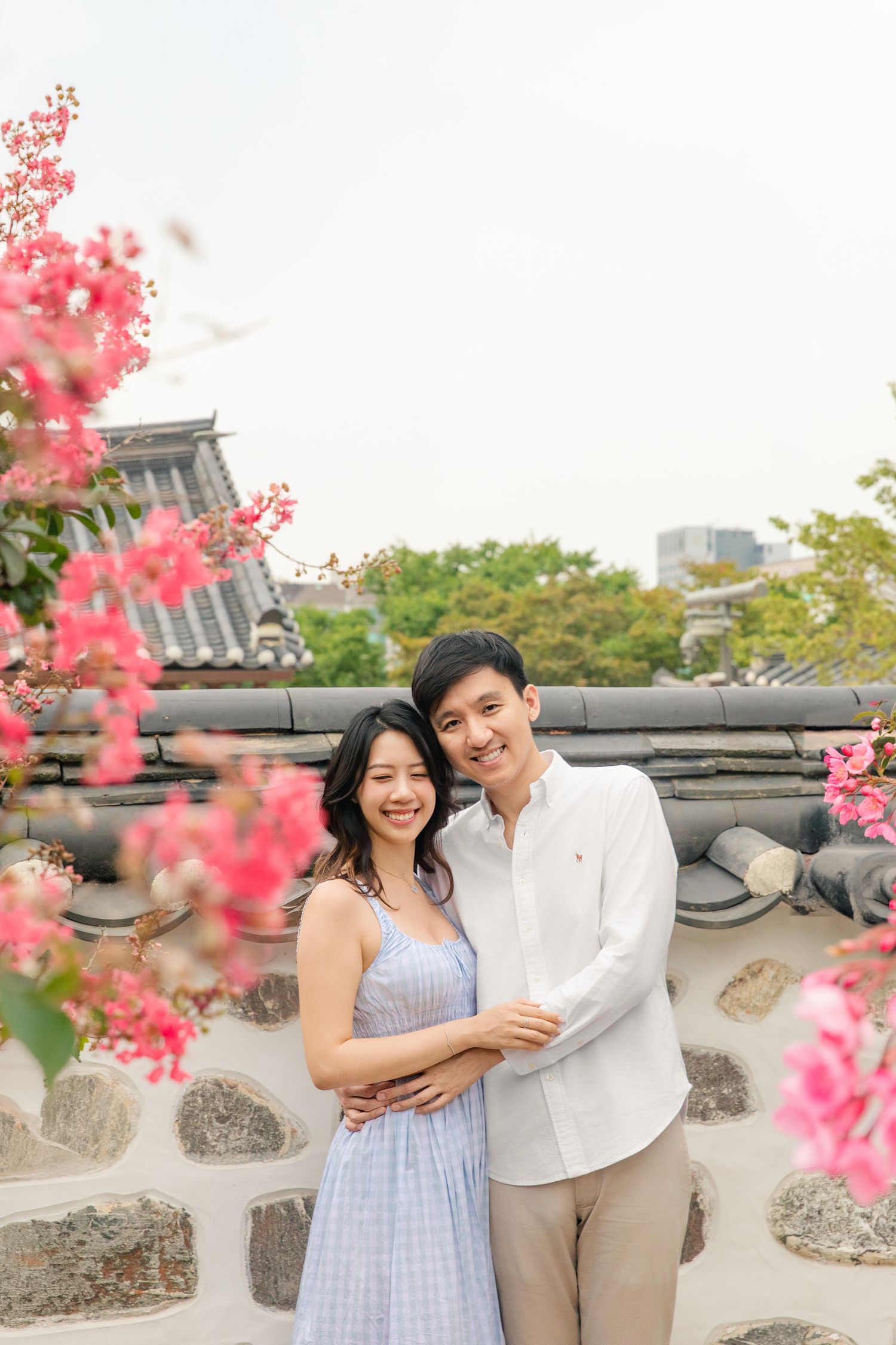 A couple shares a romantic moment near pink flowering trees at a historic stone wall.