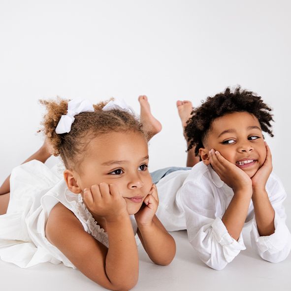 Two young children in white outfits lying on their stomachs and resting their faces on their hands while smiling.