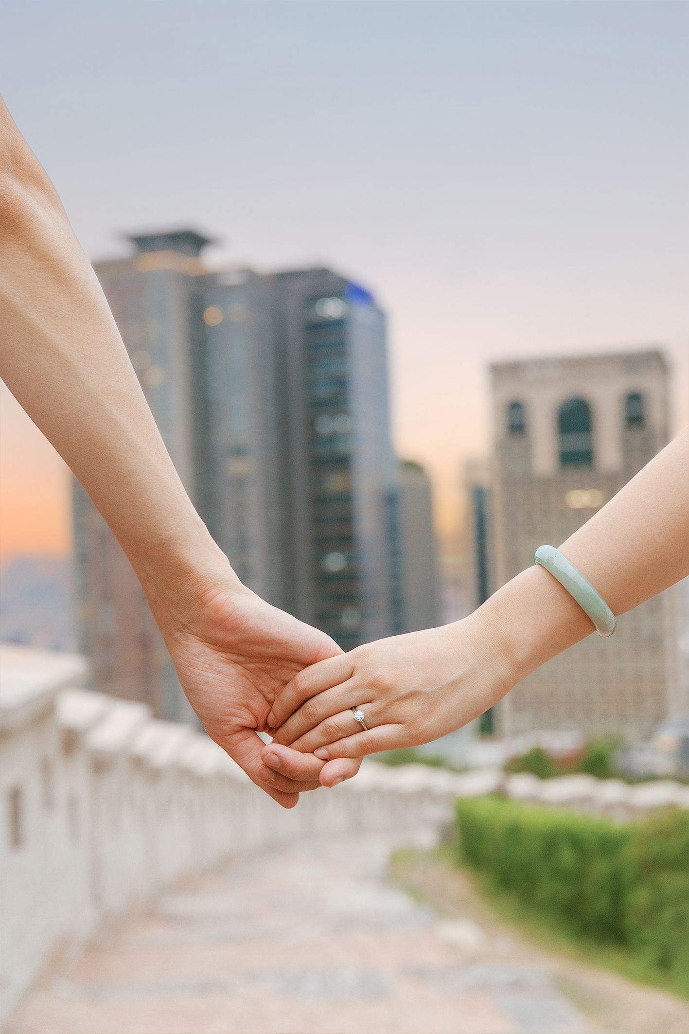Couple holding hands with a jade bracelet and ring against a blurred cityscape at sunset.