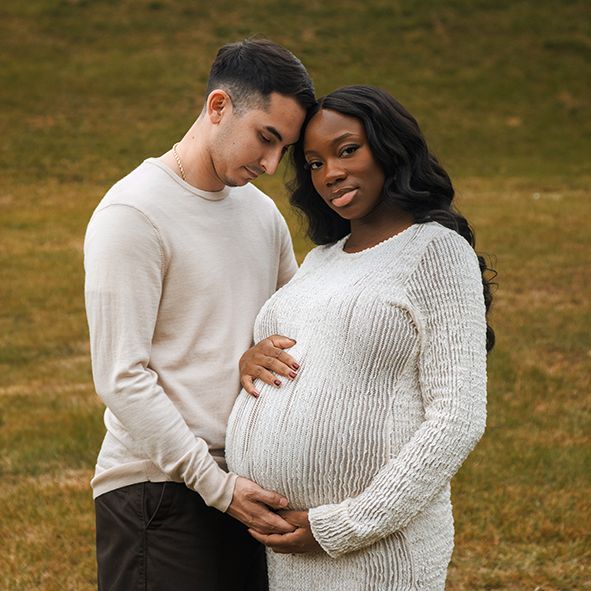 A couple in cozy white sweaters share an intimate moment outdoors during a maternity photoshoot.