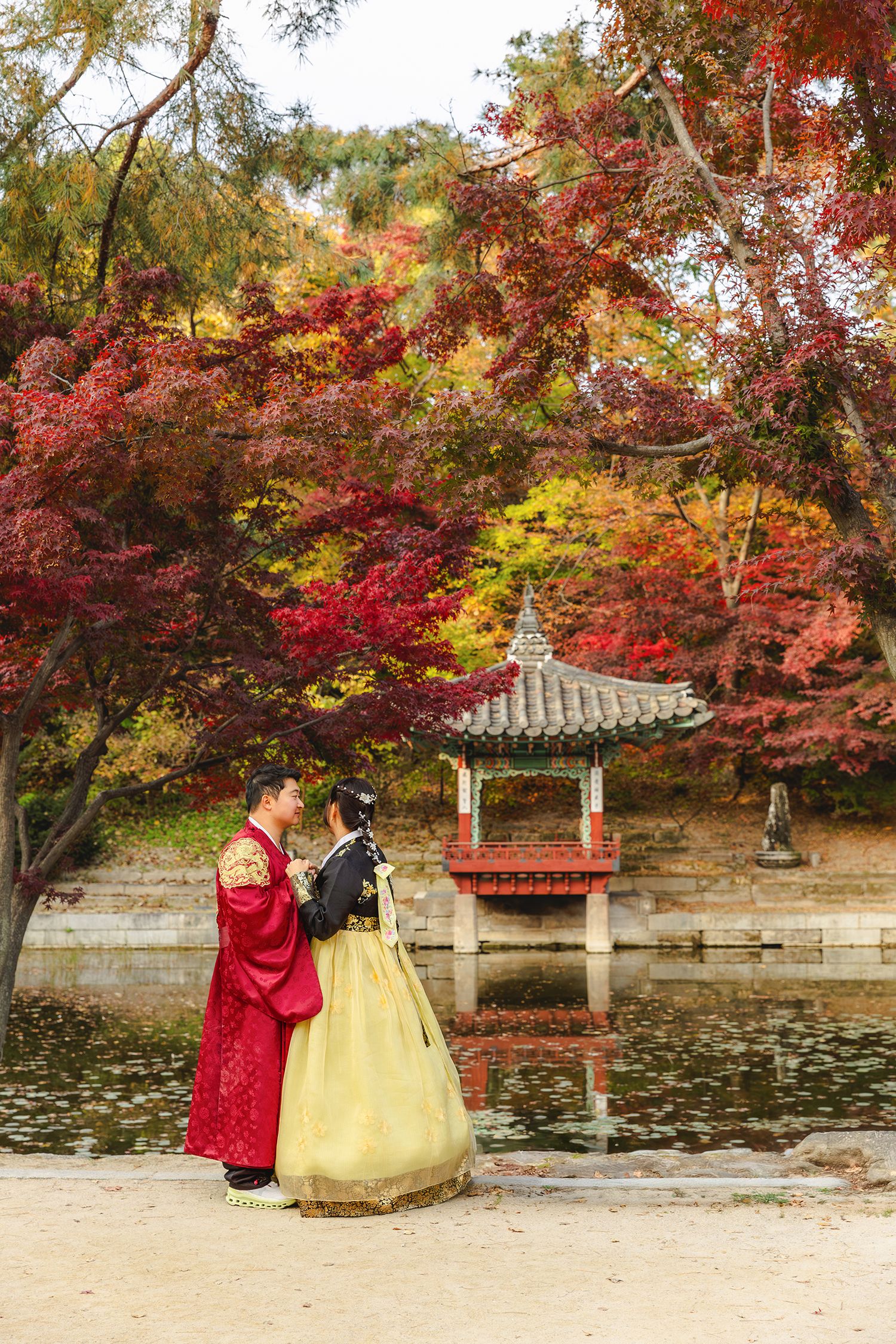 Traditional Korean hanbok outfits worn at a lakeside pavilion surrounded by vibrant red and yellow autumn foliage.