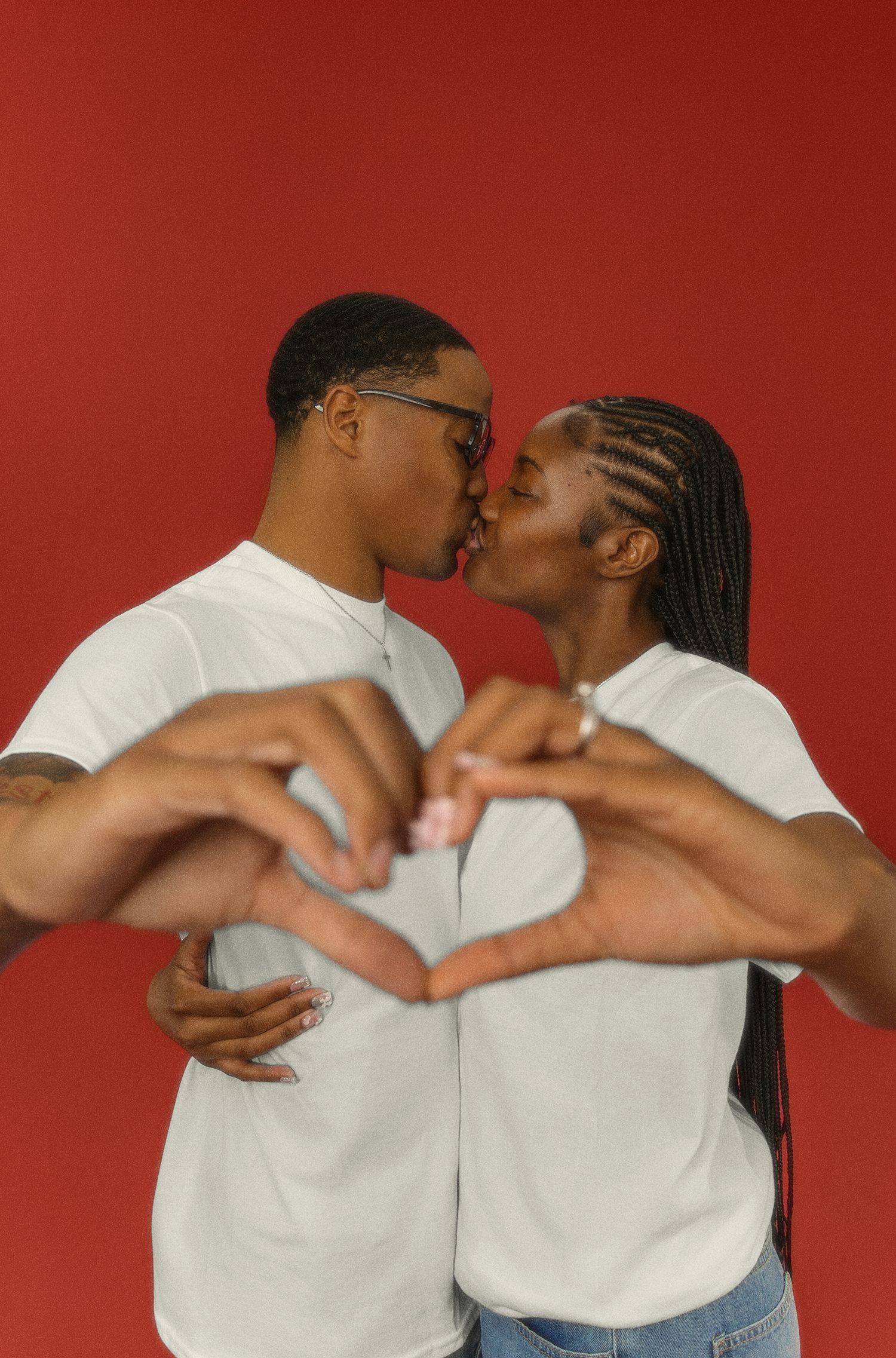 A couple in white shirts forms heart shapes with their arms while sharing an intimate moment against a red background.