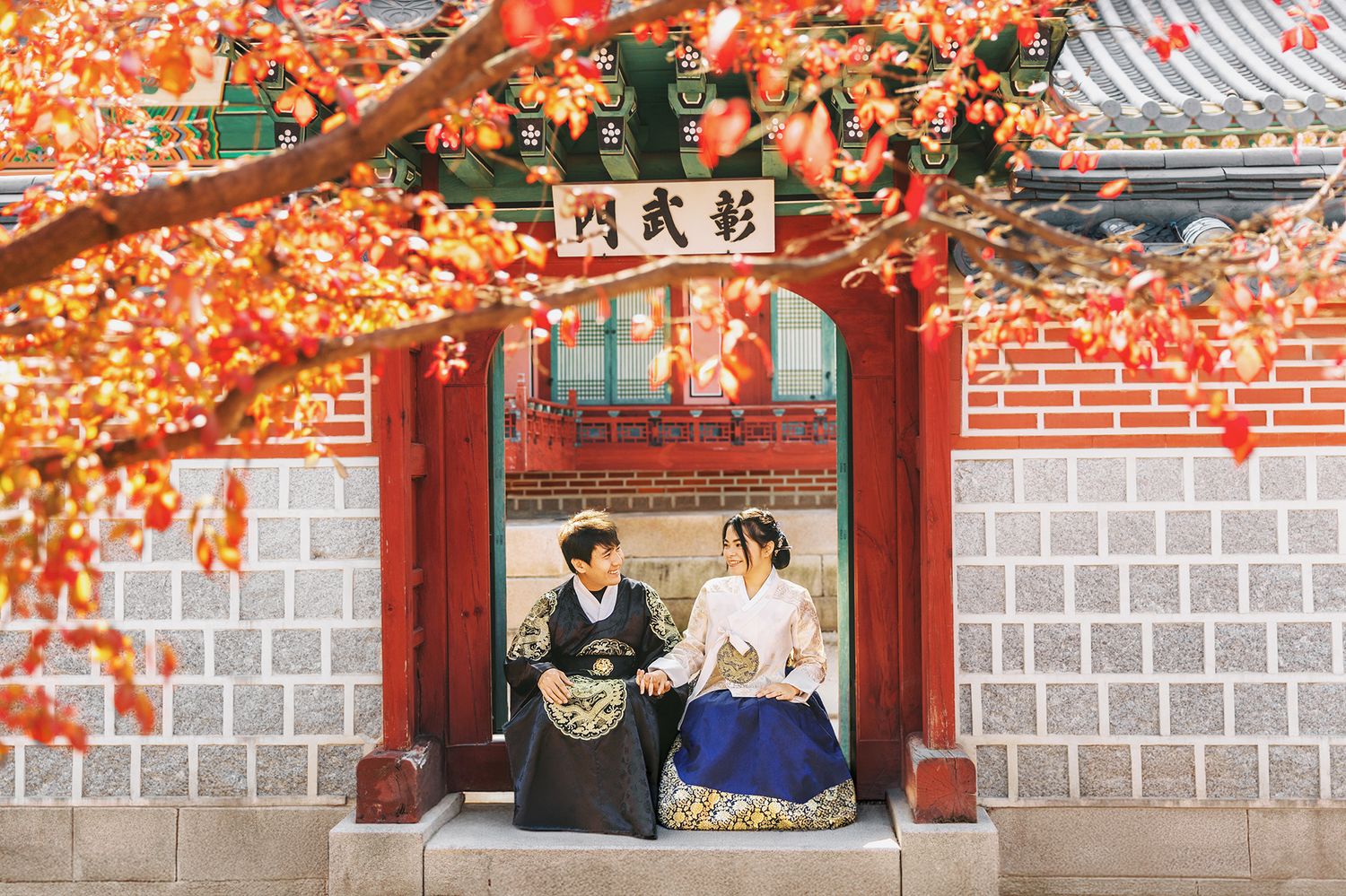 Two people in traditional Korean hanbok clothing sit beneath autumn maple leaves at a traditional palace gate in Seoul.