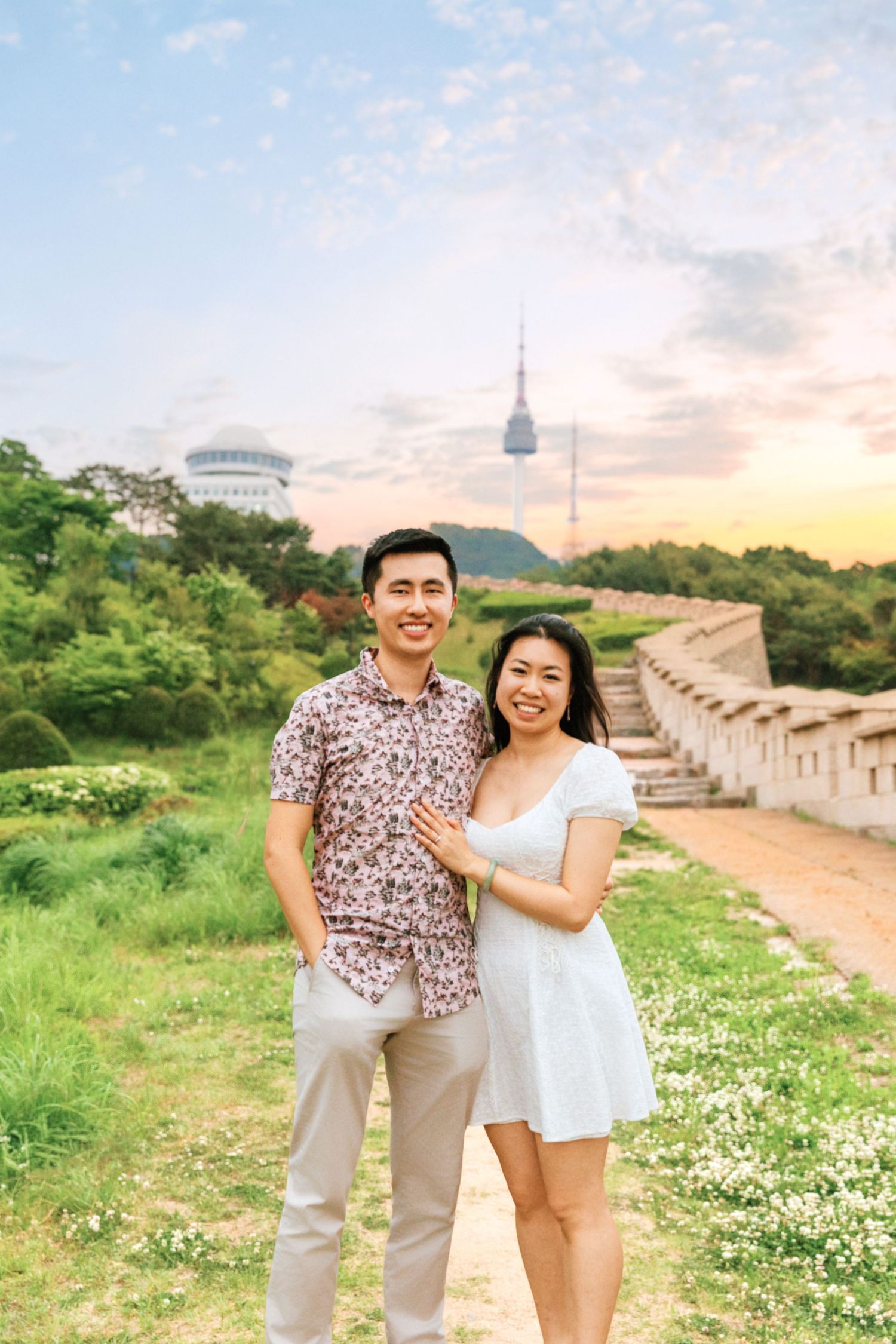 Couple posing together at sunset near Namsan Tower in Seoul, South Korea, with lush greenery and historic walls.