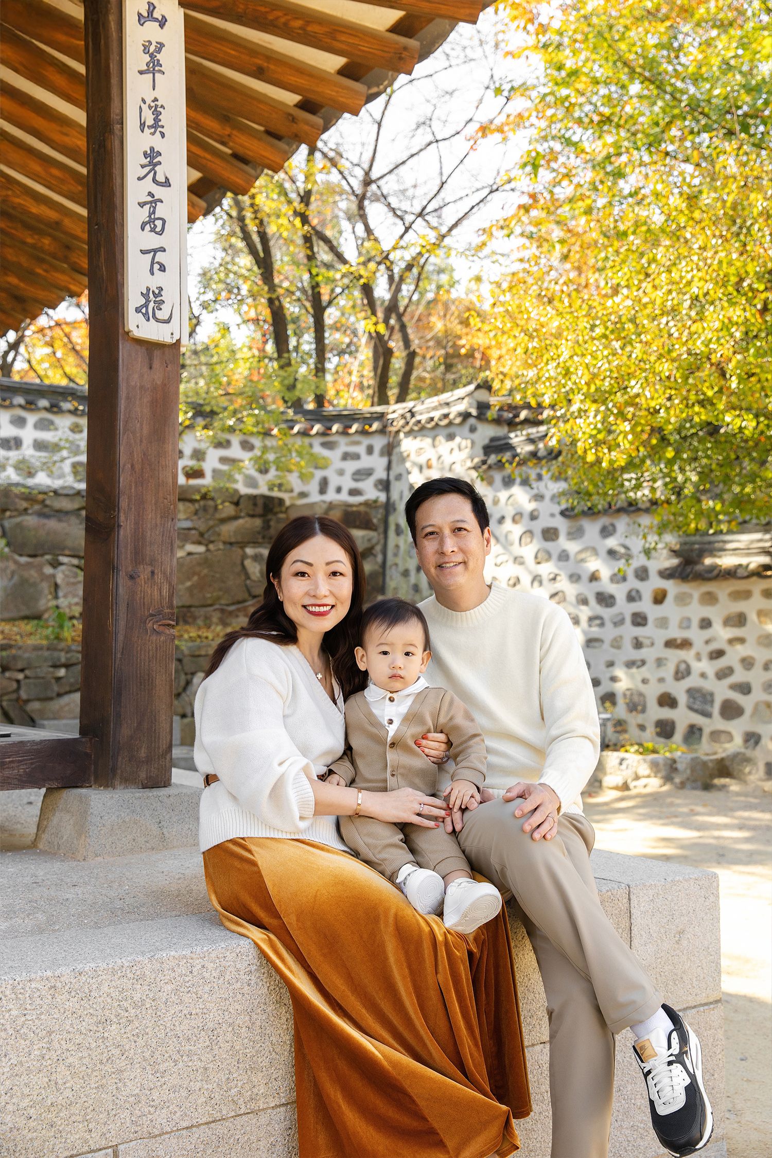 A family sits together on stone steps in a traditional Asian garden setting with autumn foliage in the background.