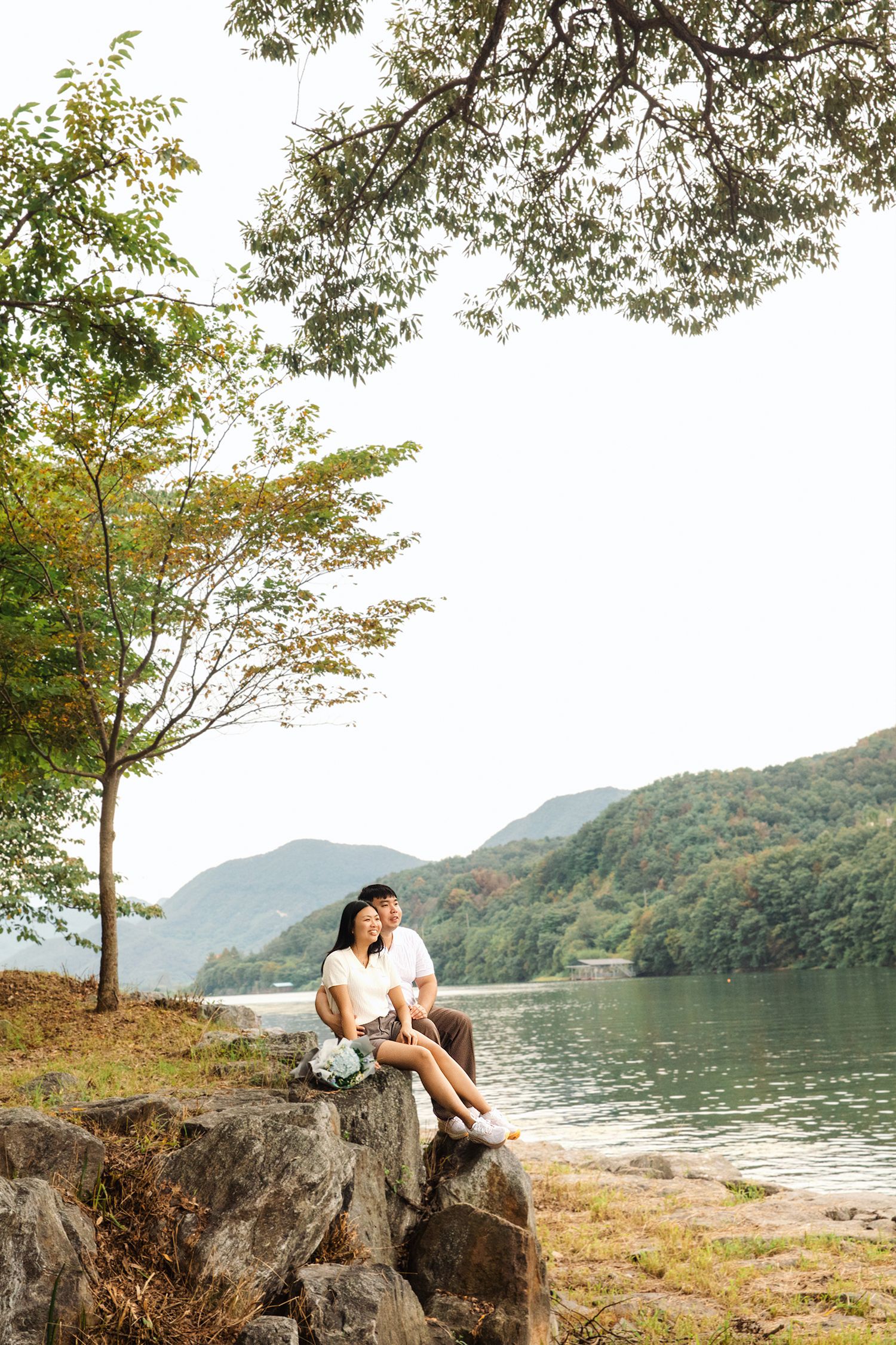 A person sits peacefully on rocks by a serene mountain lake surrounded by autumn trees and misty hills.