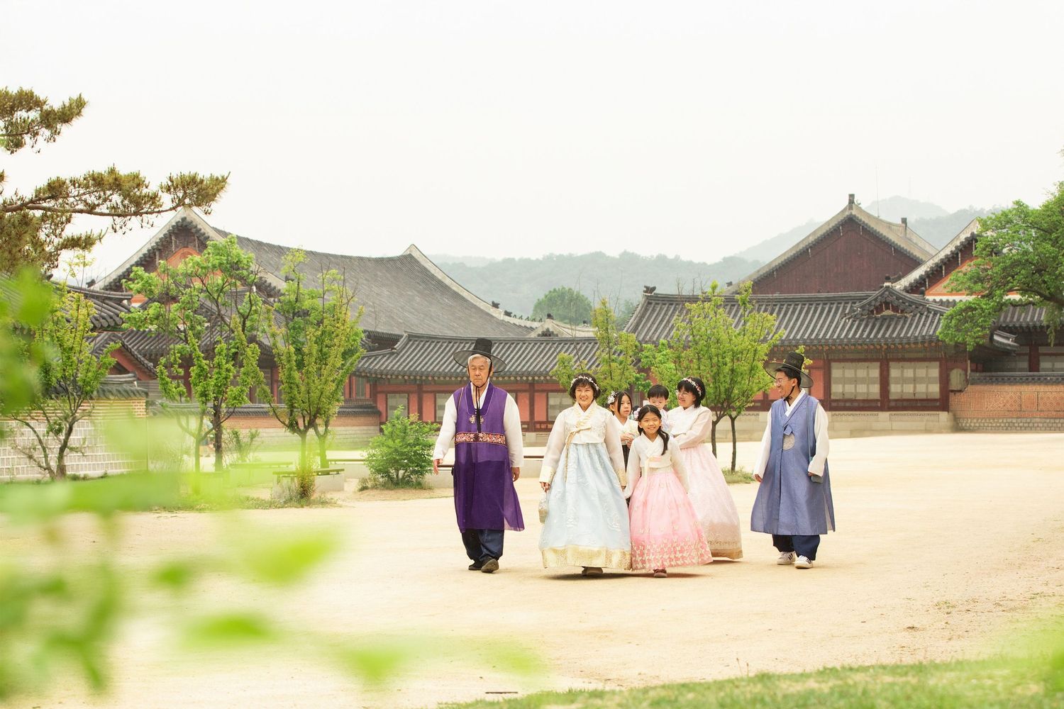 People wearing traditional Korean hanbok clothing walk along paths between historic palace buildings in Seoul.