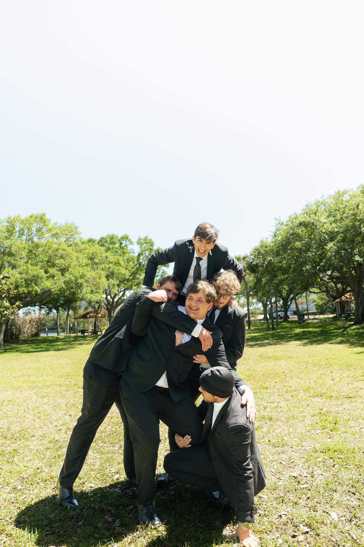 Groomsmen in black suits playfully posing together outdoors in a park on a sunny wedding day.