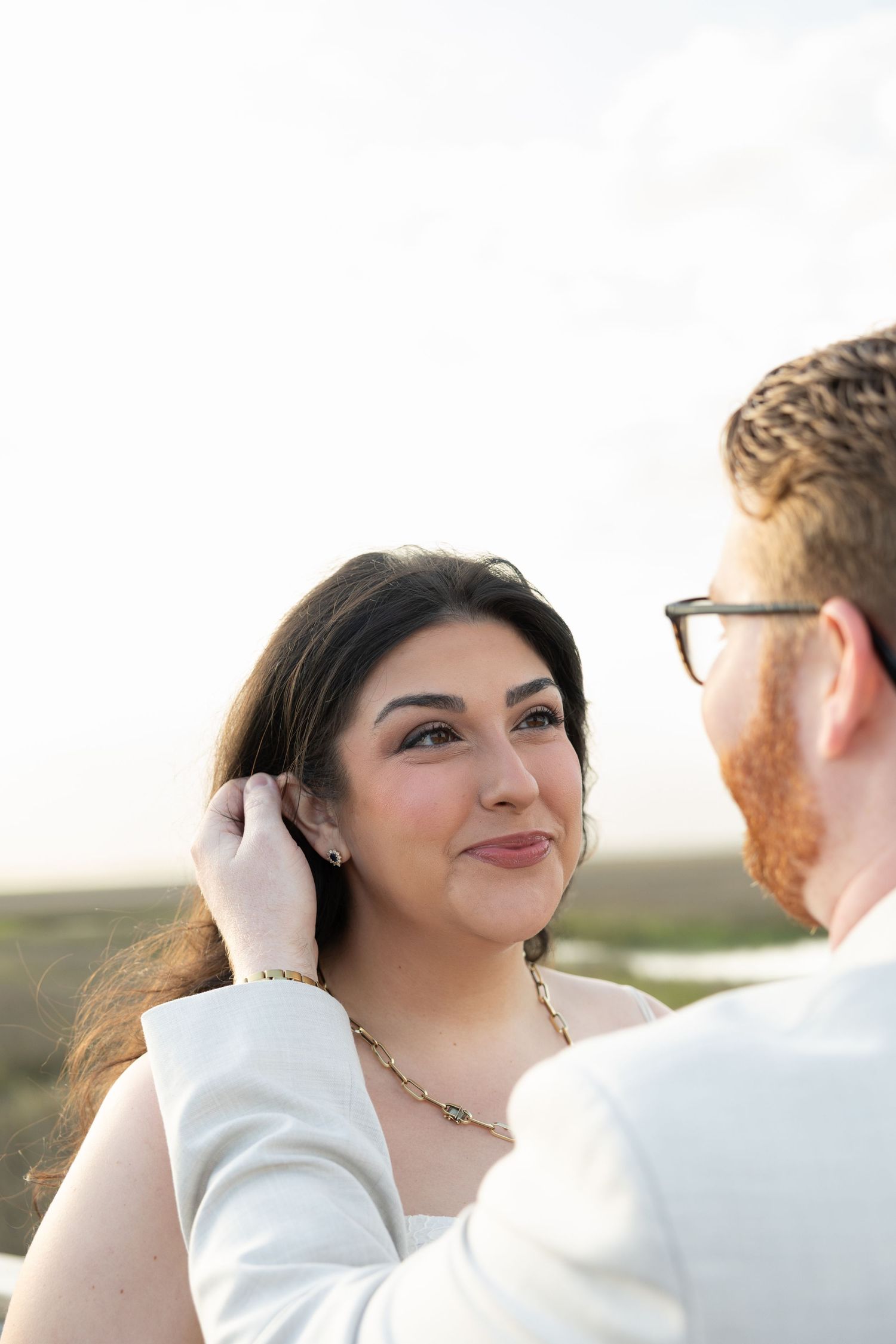 Bride smiling tenderly as groom gently tucks her dark hair back during an outdoor wedding portrait.