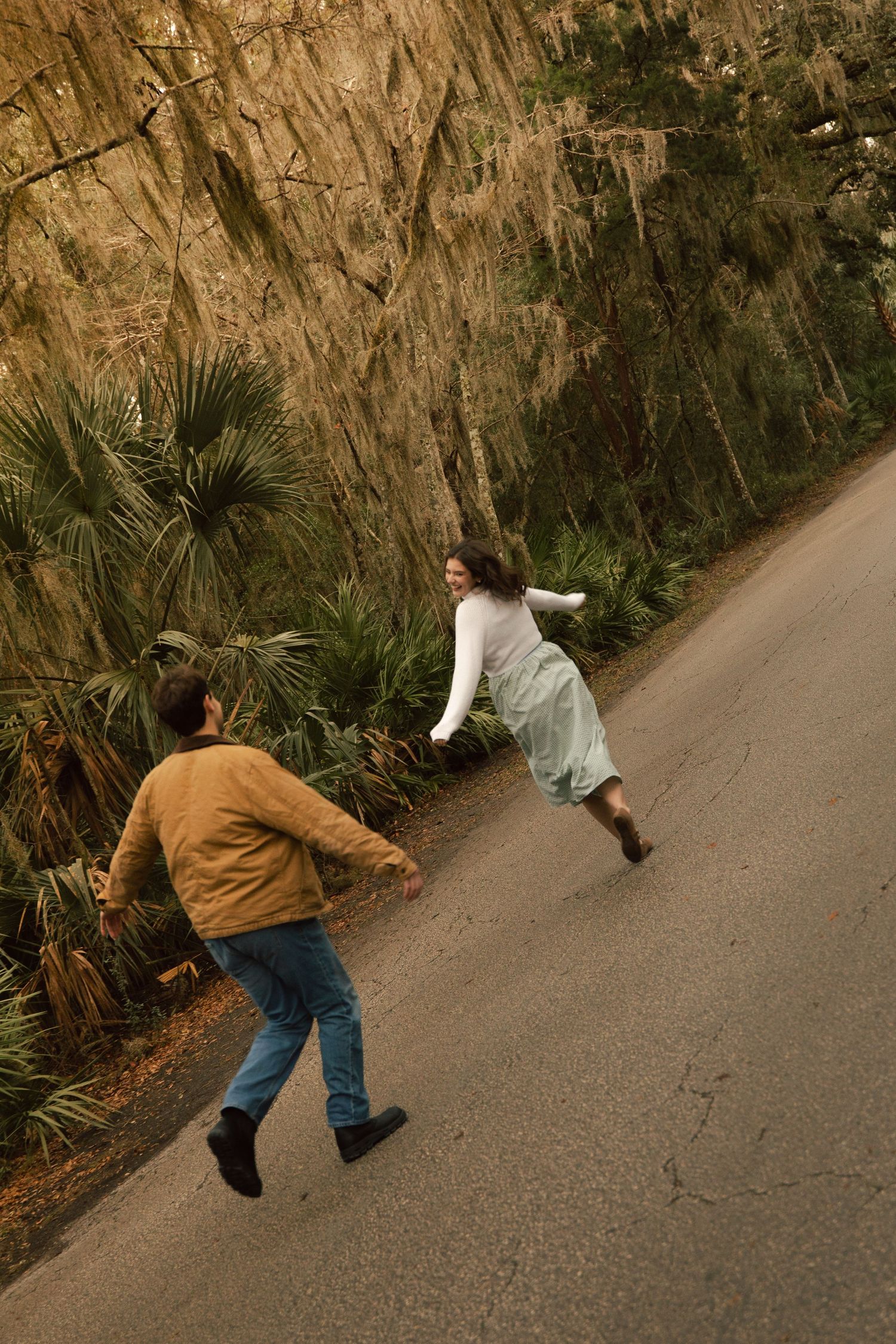 Two people playfully running on a winding road lined with palm trees and rocky cliffs.