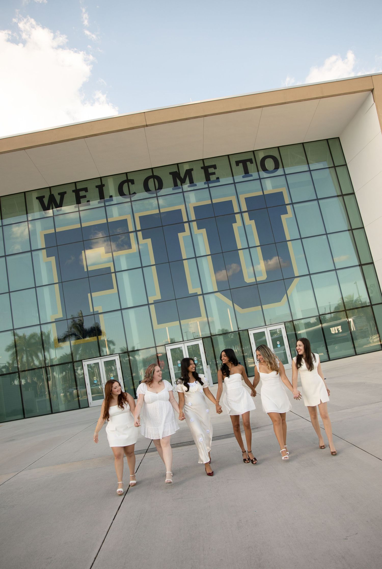 A group in white dresses walks along the exterior of a modern glass building with a welcome sign.