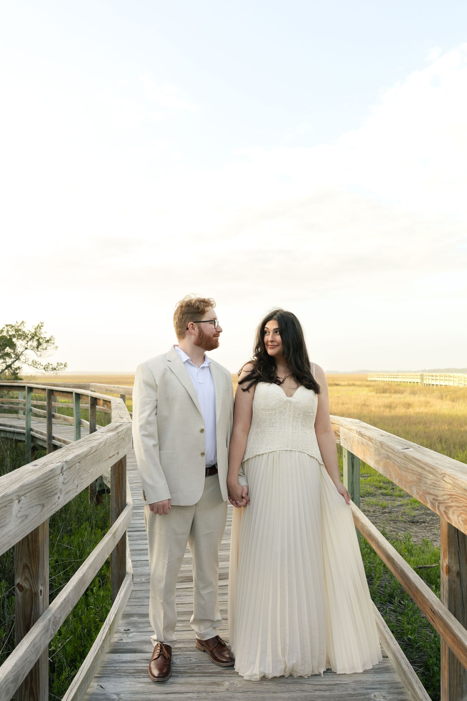 A couple in wedding attire stands on a wooden boardwalk surrounded by golden marshlands at sunset.