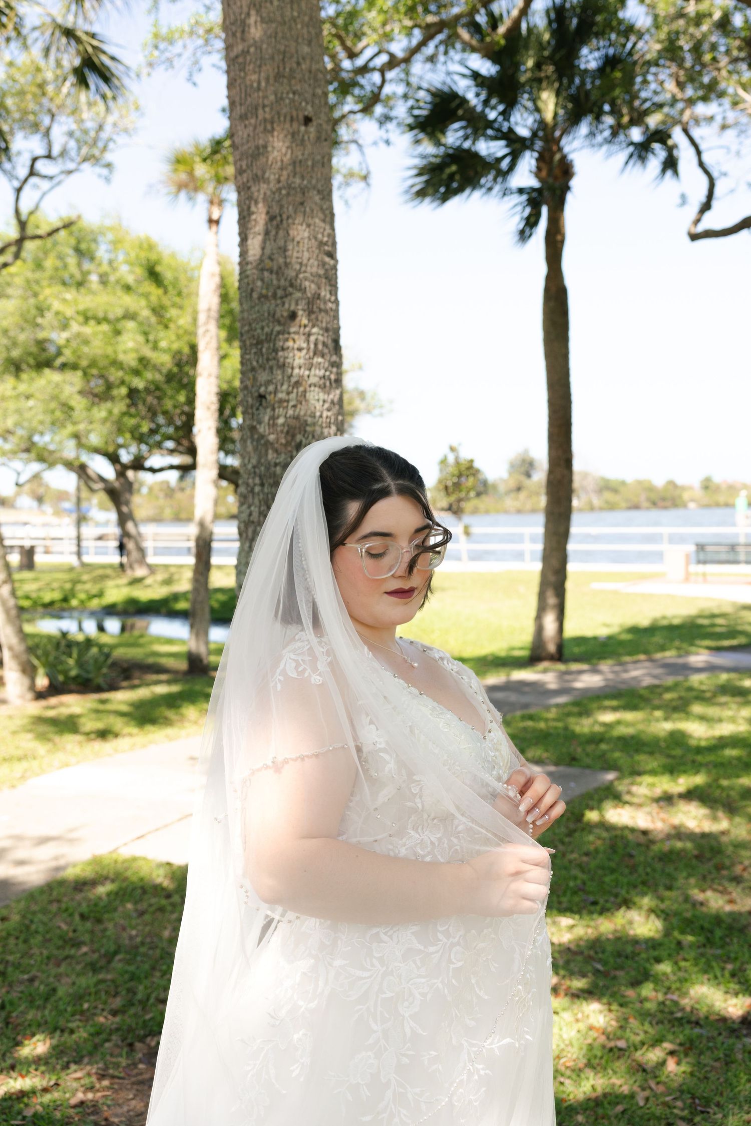 Bride in lace wedding gown and veil poses outdoors near palm trees by waterfront on sunny day.