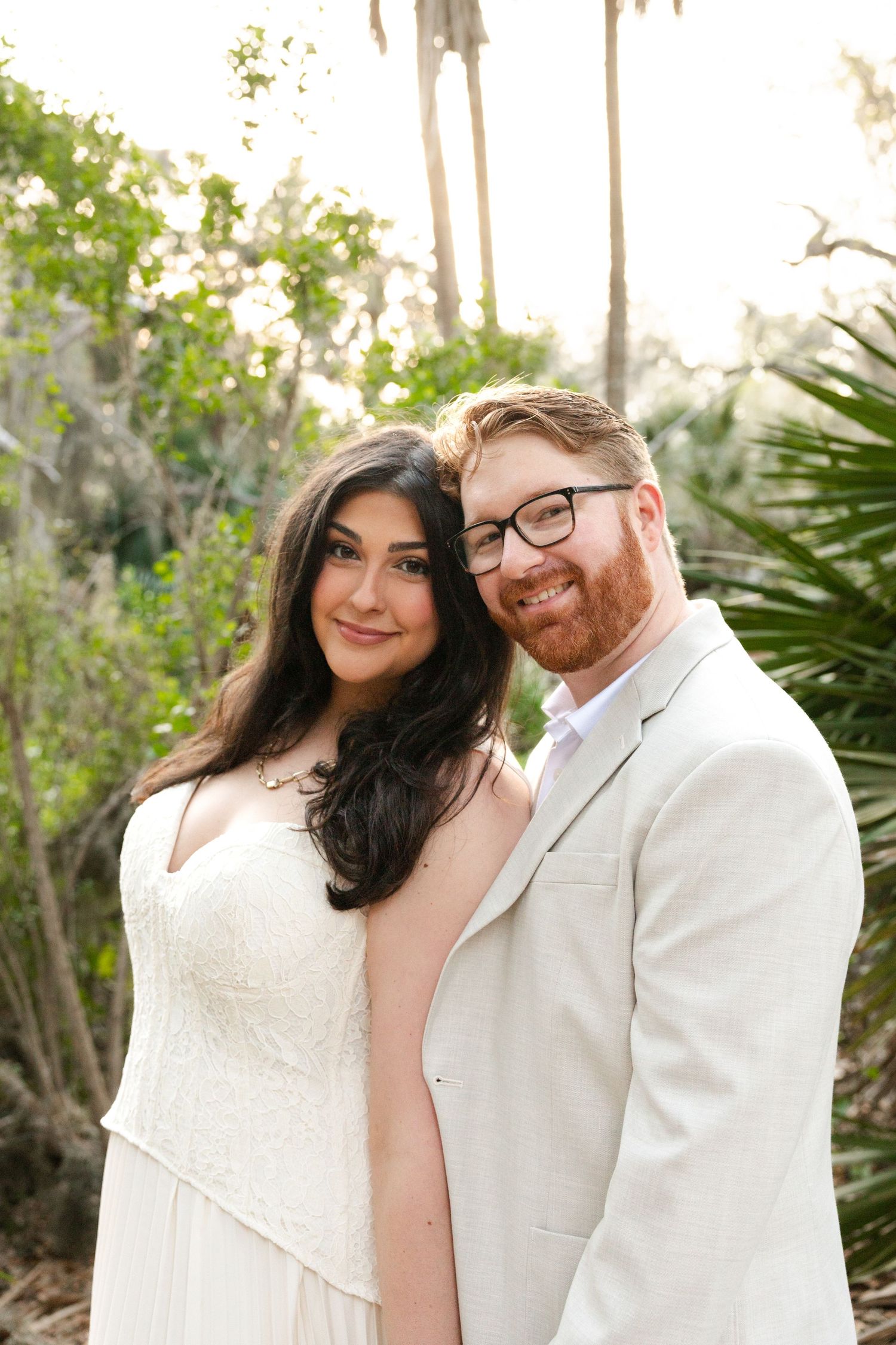 A smiling couple in wedding attire poses outdoors among lush greenery, the bride in white lace and groom in a tan suit.