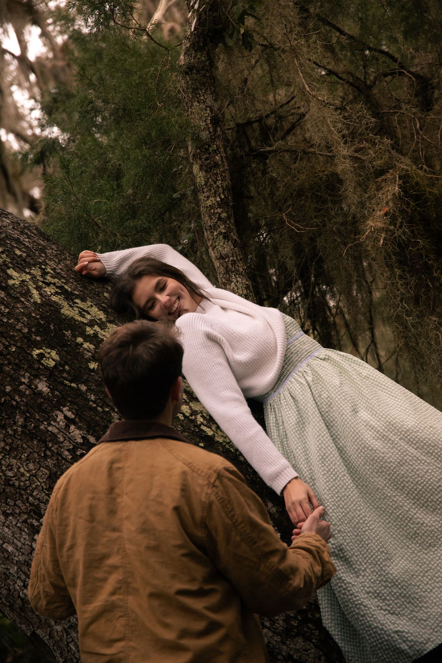 A person in a white dress and brown jacket lies peacefully against a tree trunk in a moody forest setting.