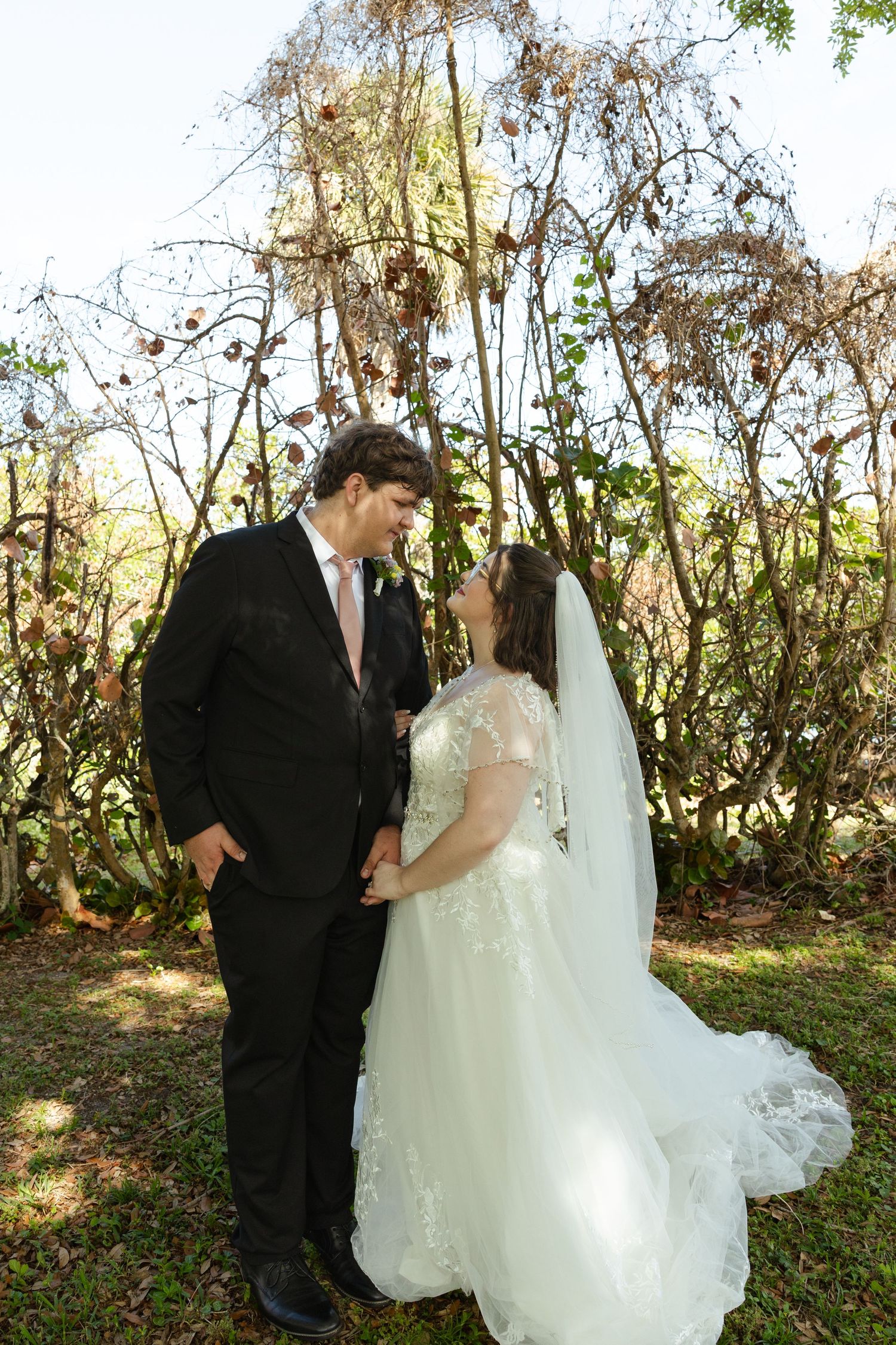 Bride and groom share a romantic moment outdoors, with the bride in a white lace gown and veil, groom in a black suit.
