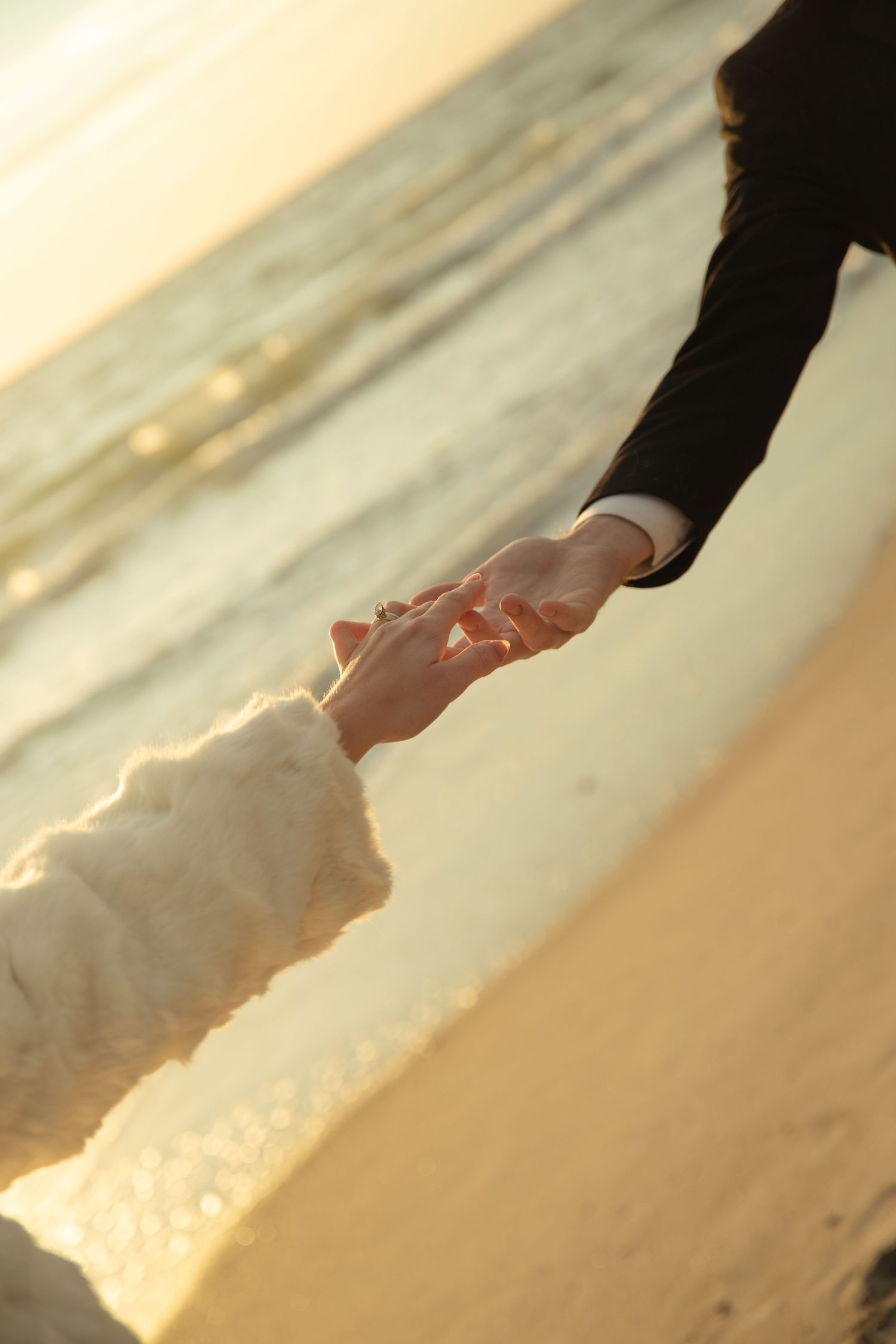 Couple holding hands on a sandy beach at golden sunset, wearing formal wedding attire near ocean waves.