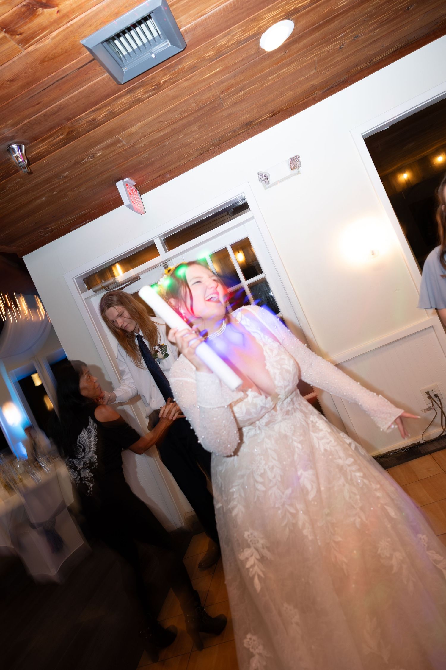 Wedding guests dance and celebrate on a wooden-floored reception venue with white walls and warm lighting.