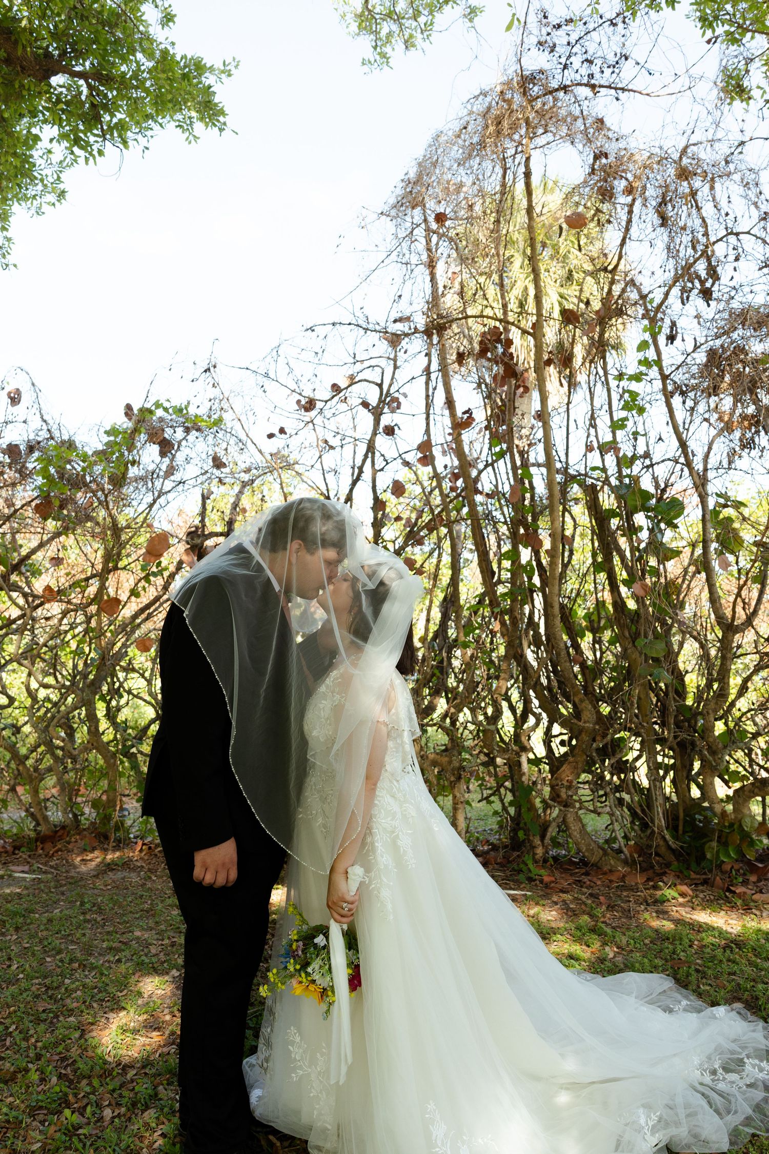 Bride and groom sharing a kiss under a wedding veil outdoors, surrounded by lush greenery and rustic branches.