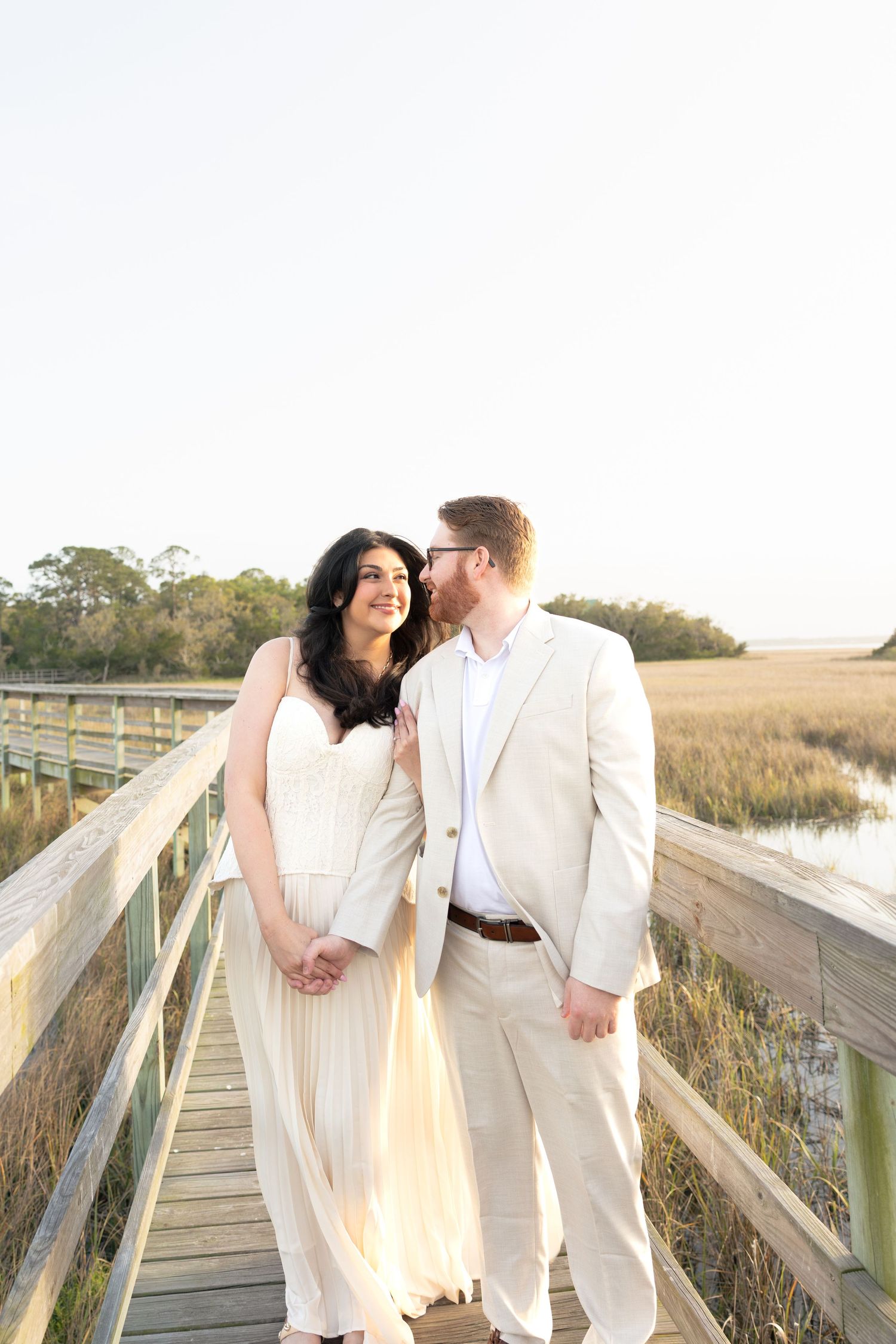 Couple in white attire sharing a romantic moment on a wooden boardwalk surrounded by golden marshlands.