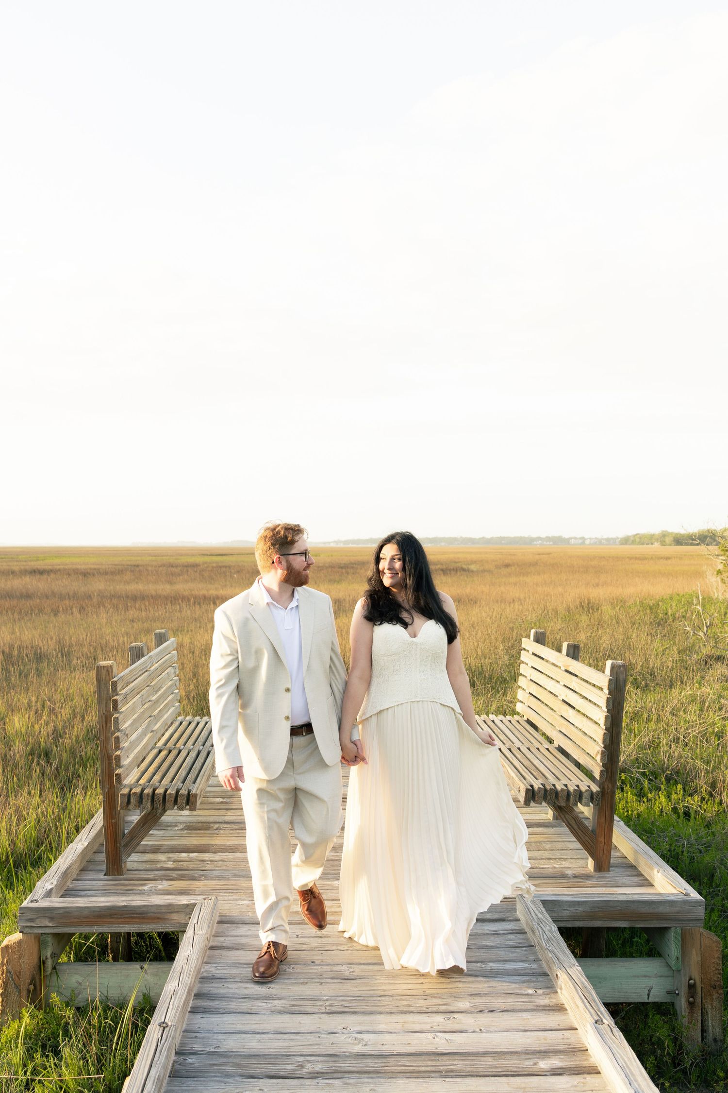 A bride and groom walk hand-in-hand on a wooden boardwalk surrounded by golden marshlands at sunset.