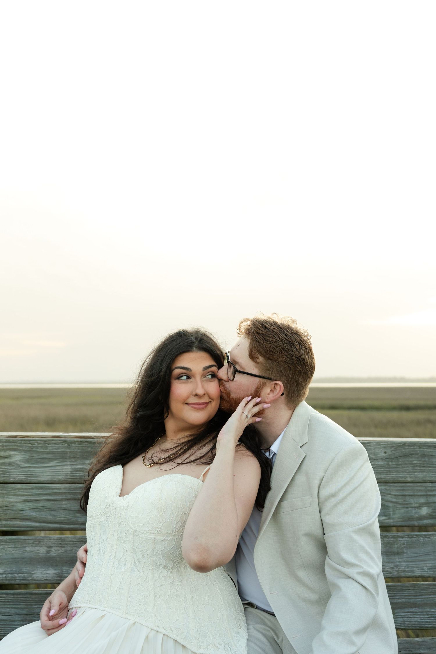 A groom in a tan suit whispers to his smiling bride in a white dress on a wooden bench by the shore.