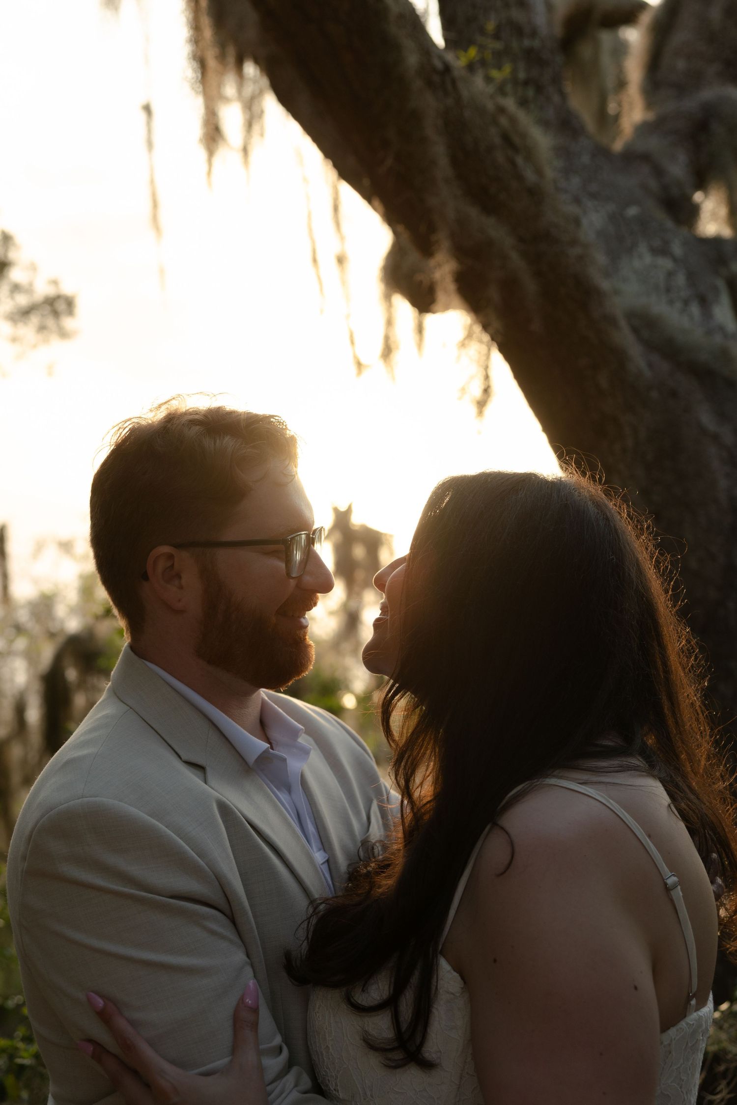 Couple shares romantic moment under Spanish moss-draped oak tree, bathed in golden sunset light.