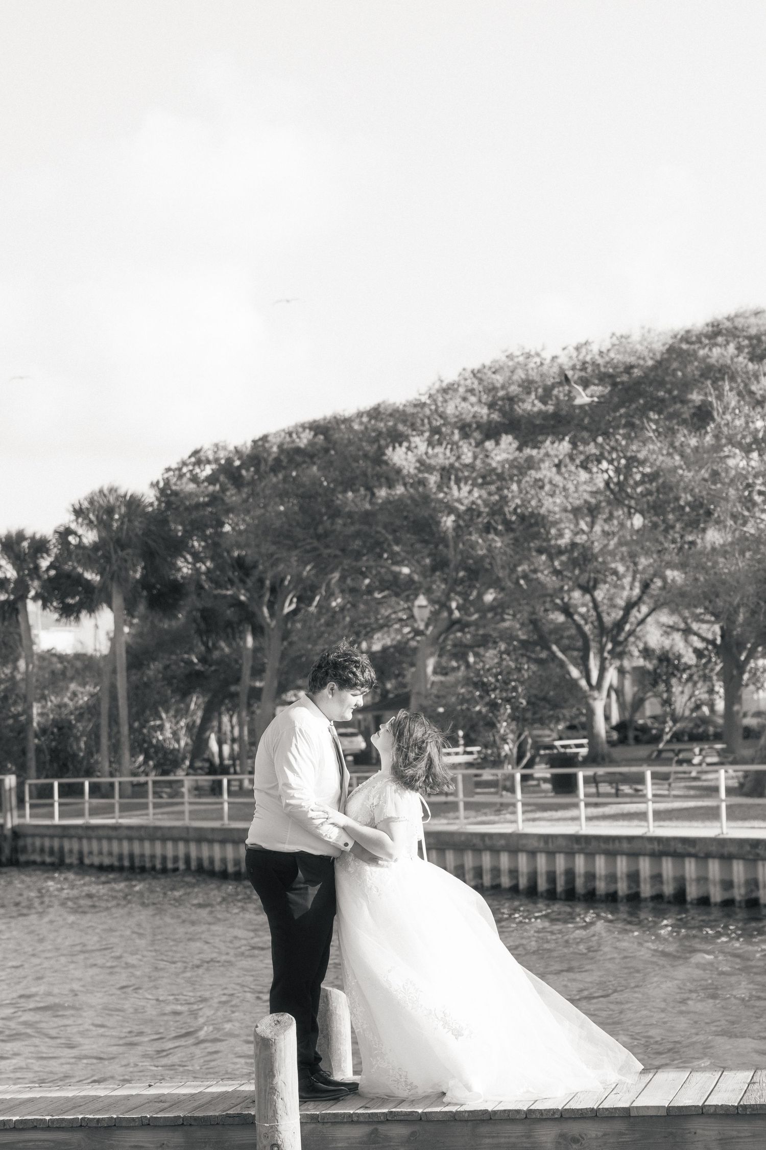 Black and white photo of a bride and groom sharing a romantic moment on a waterfront dock surrounded by lush trees.