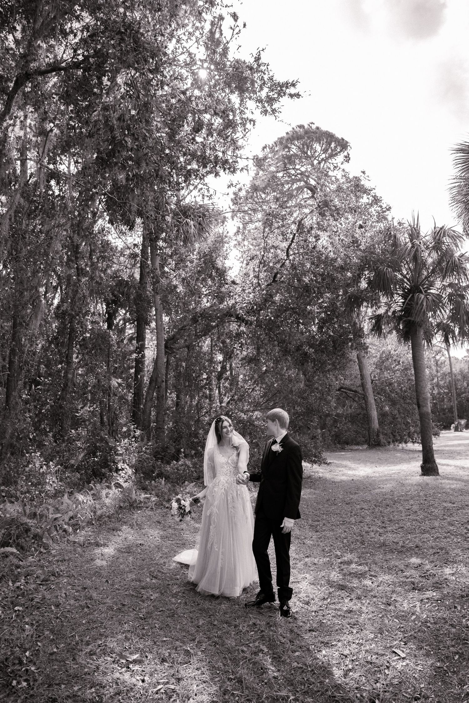 Black and white photo of a couple in wedding attire walking along a tree-lined path.