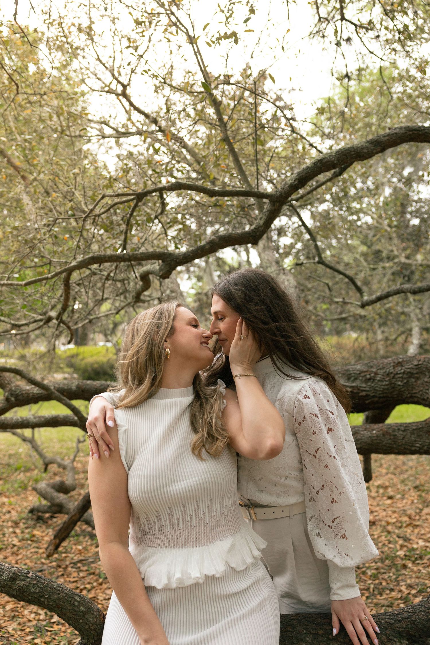Two people share an intimate moment under a large tree branch in a park setting during autumn.