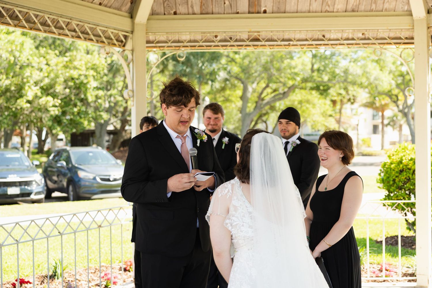 Outdoor wedding ceremony in a gazebo with officiant reading vows to bride in white gown and veil.