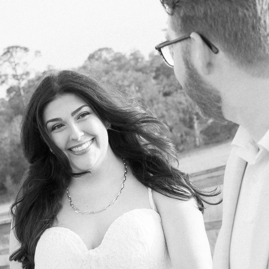 Black and white photo of a smiling bride in a strapless gown looking at a man in glasses outdoors.